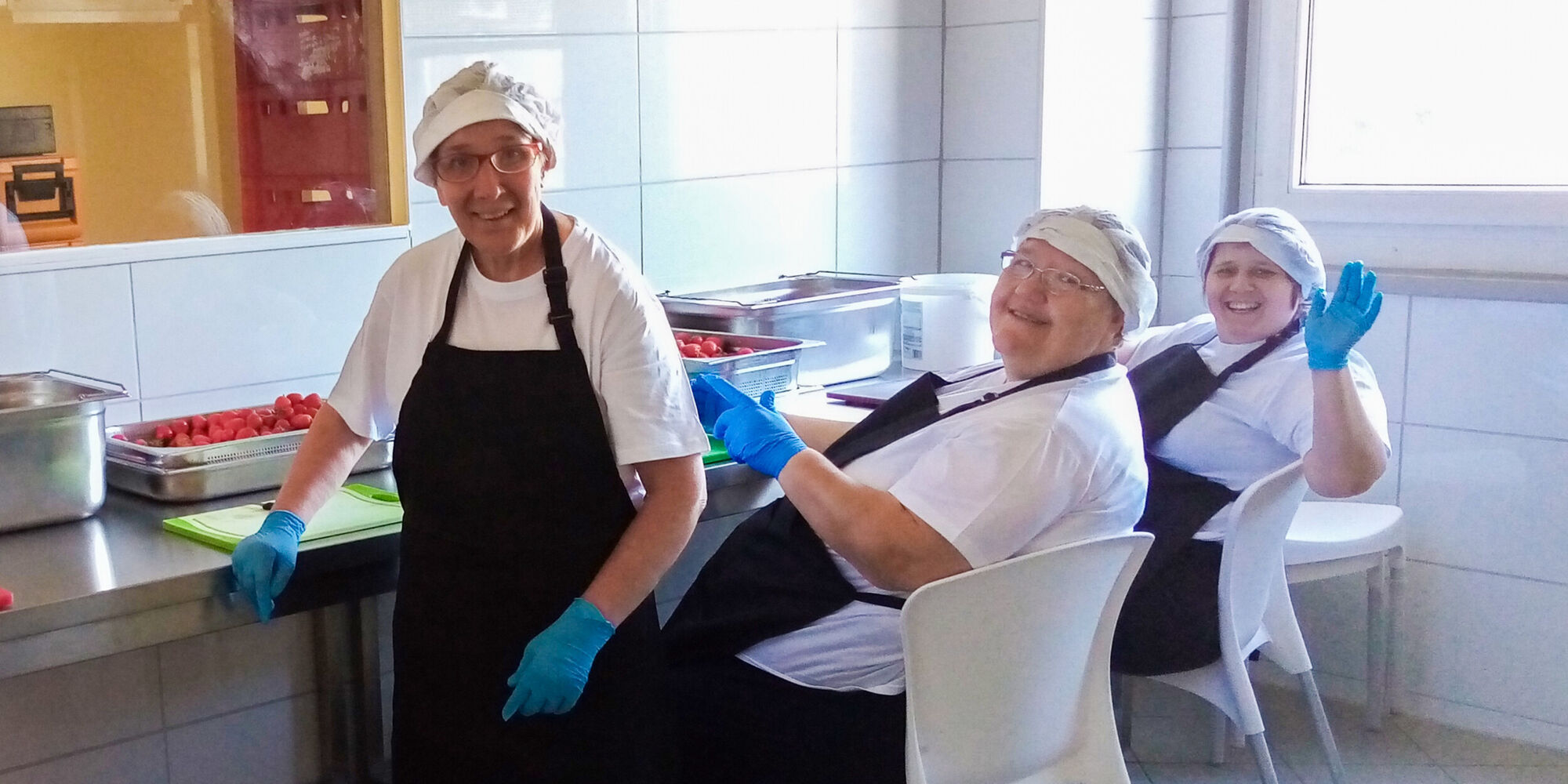 Vier Menschen mit Kochsch&uuml;rze und Haarnetz, Gummihandschuhen winken in die Kamera.
