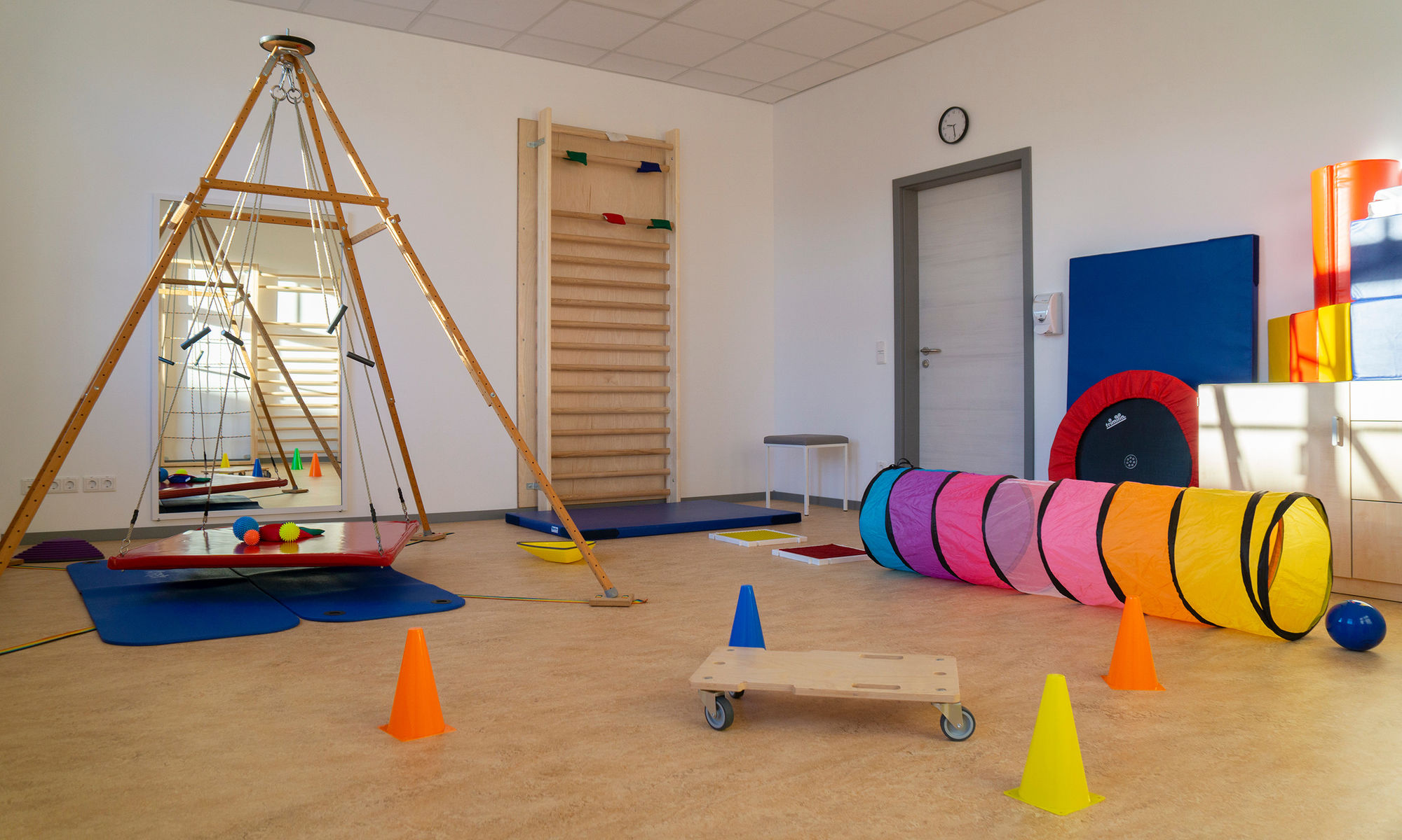 ein großer Raum mit Therapiematerialien wie zum Beispiel Matten, Sprossenwand, Schaukel, Kegeln, Bällen