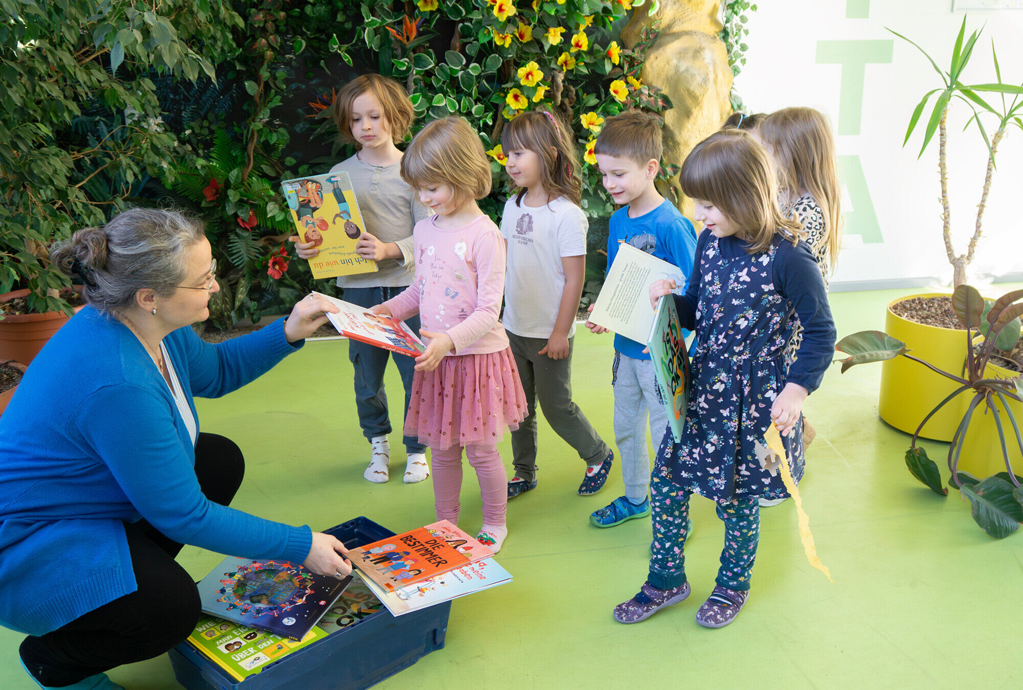 Ein l&auml;chelnde Frau hockt vor einer Kiste voller B&uuml;cher und &uuml;bergibt diese an lachende Kita-Kinder