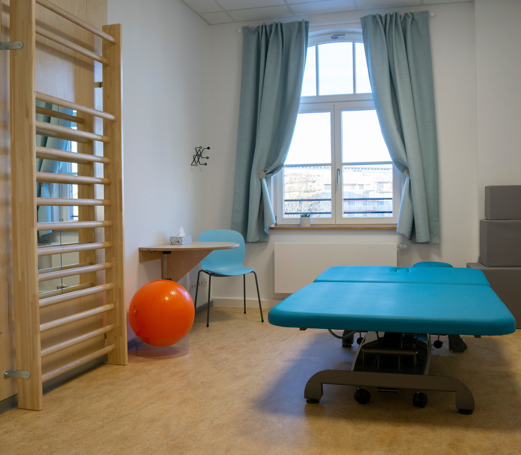 ein Therapieraum mit großer Liege, Sprossenwand, Tisch, Stuhl und Spiegel