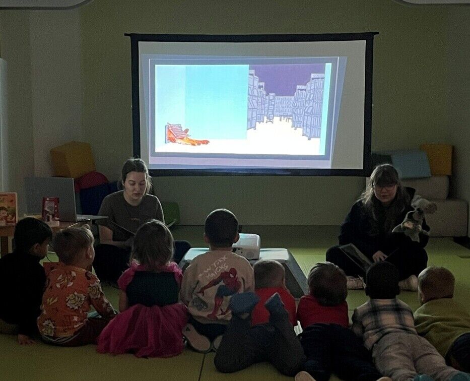 Zwei Frauen sitzen vor einem gro&szlig;en Bildschirm und schauen zu vielen Kindern. Eine Frau liest ein Buch vor, die andere hat eine Handpuppe in der Hand.