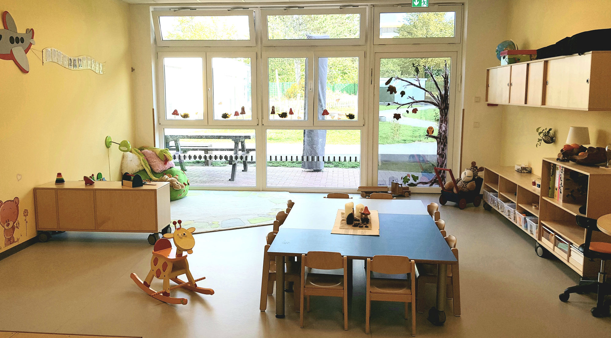 Ein Gruppenraum mit  Spielsachen, regalen, Tischen, Stühlen und bodentiefen Fenstern die zum Garten führen.