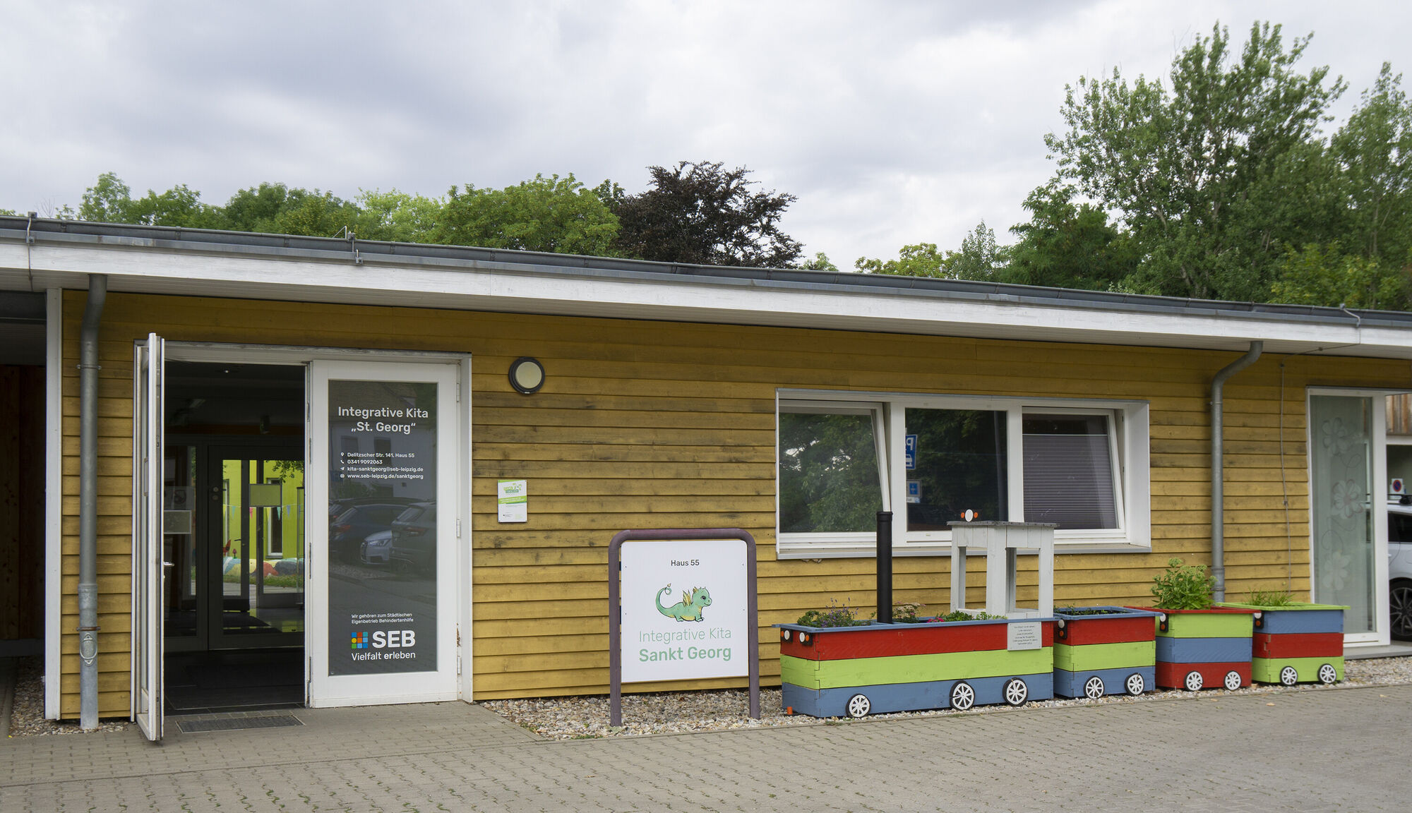 Ein flaches Gebäude aus Holz mit offener Tür. Davon sind Pflanzkübel in Form einer bunten Eisanbahn zu sehen.