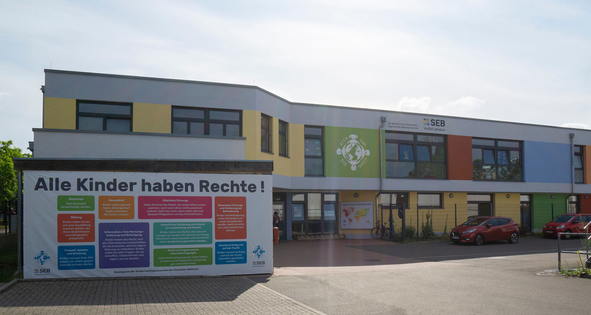 Ein großes buntes Gebäude. Davor ist ein großer Baner mit der Aufschrift "Alle Kinder haben Rechte!" zu sehen