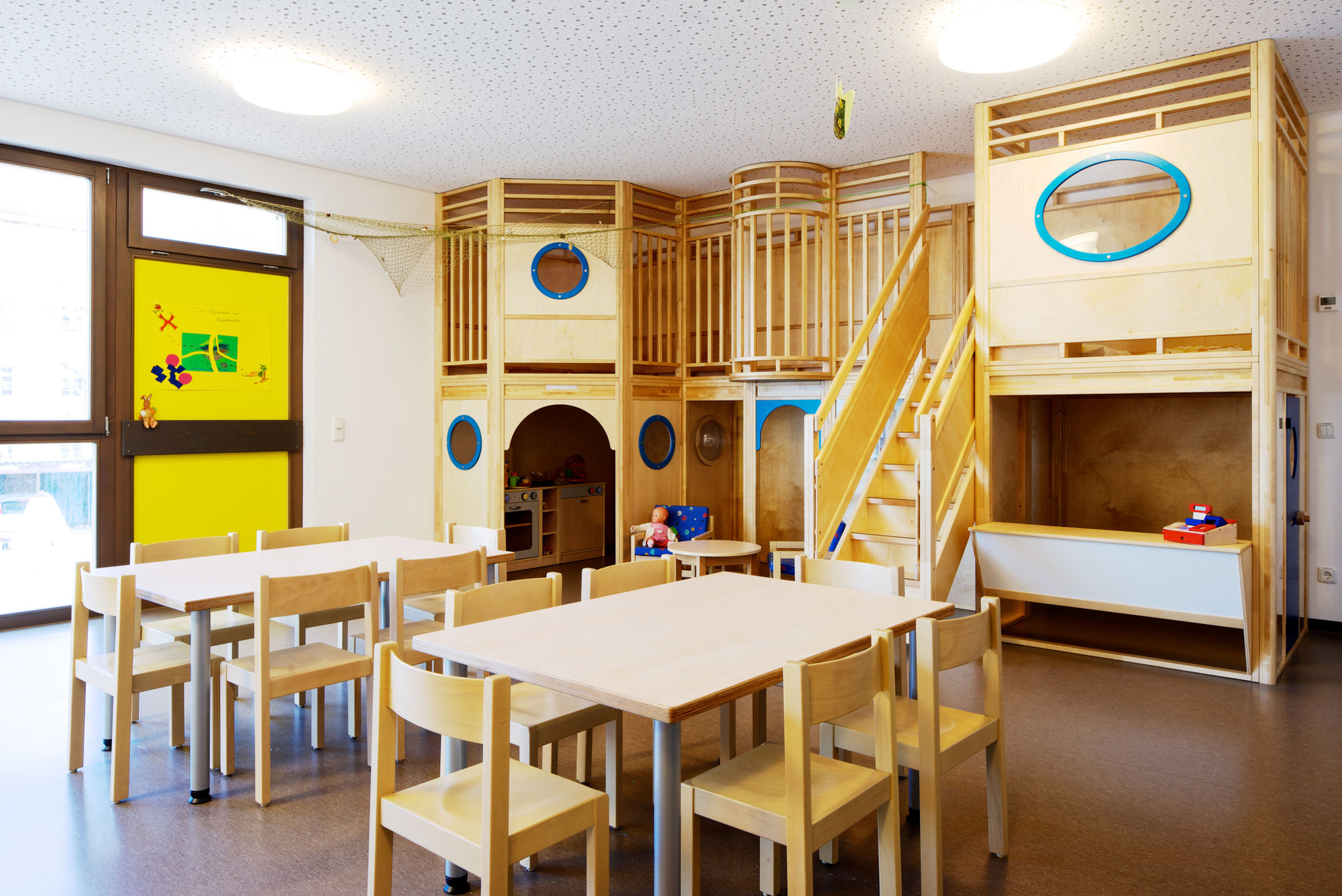 Ein heller Raum mit kleinen Tischen und Stühlen und einem eingebauten Kletterbereich aus Holz