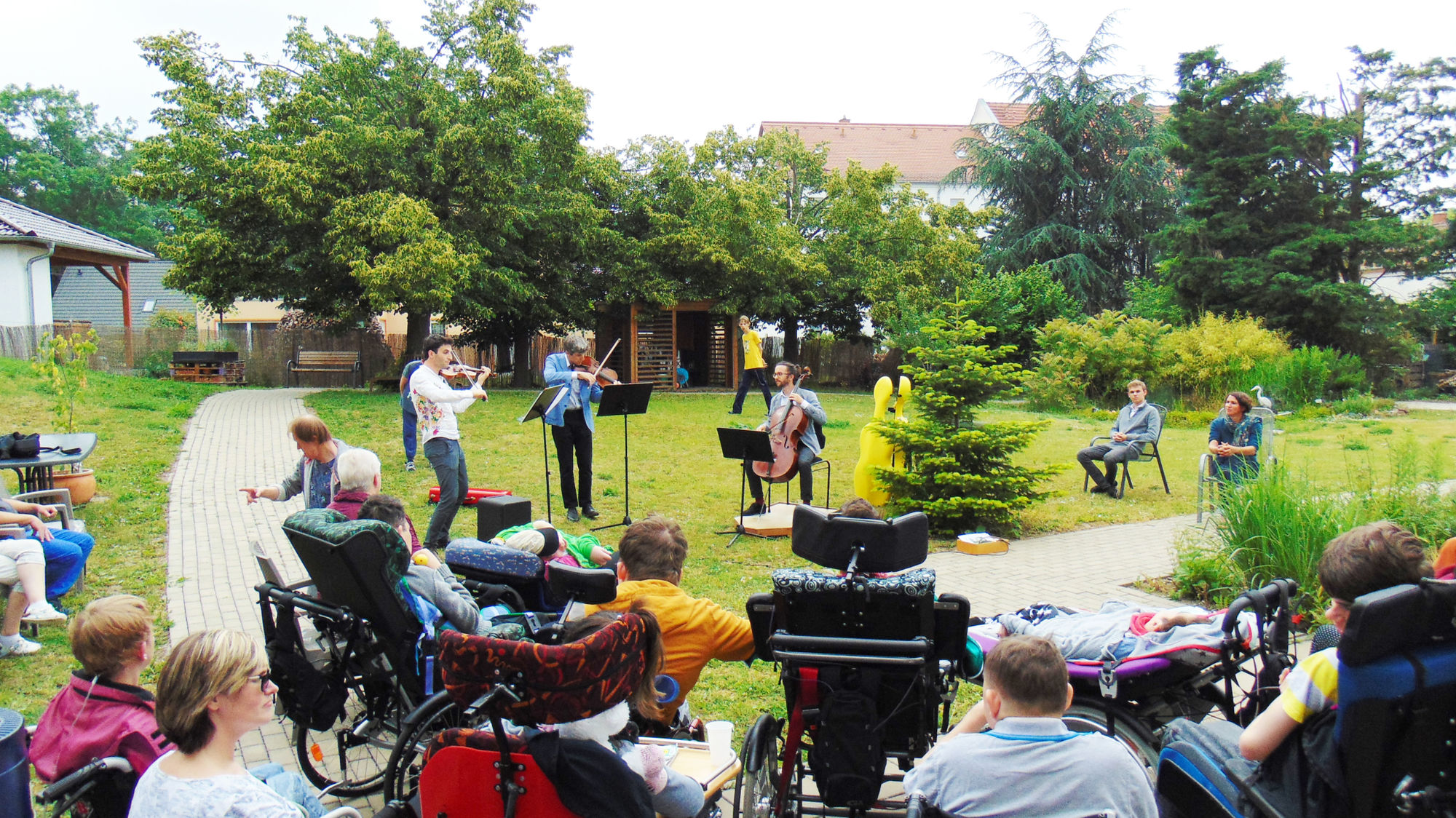Viele Menschen sind im Garten und hören und schauen einem Duo zu, welches Violine spielt.