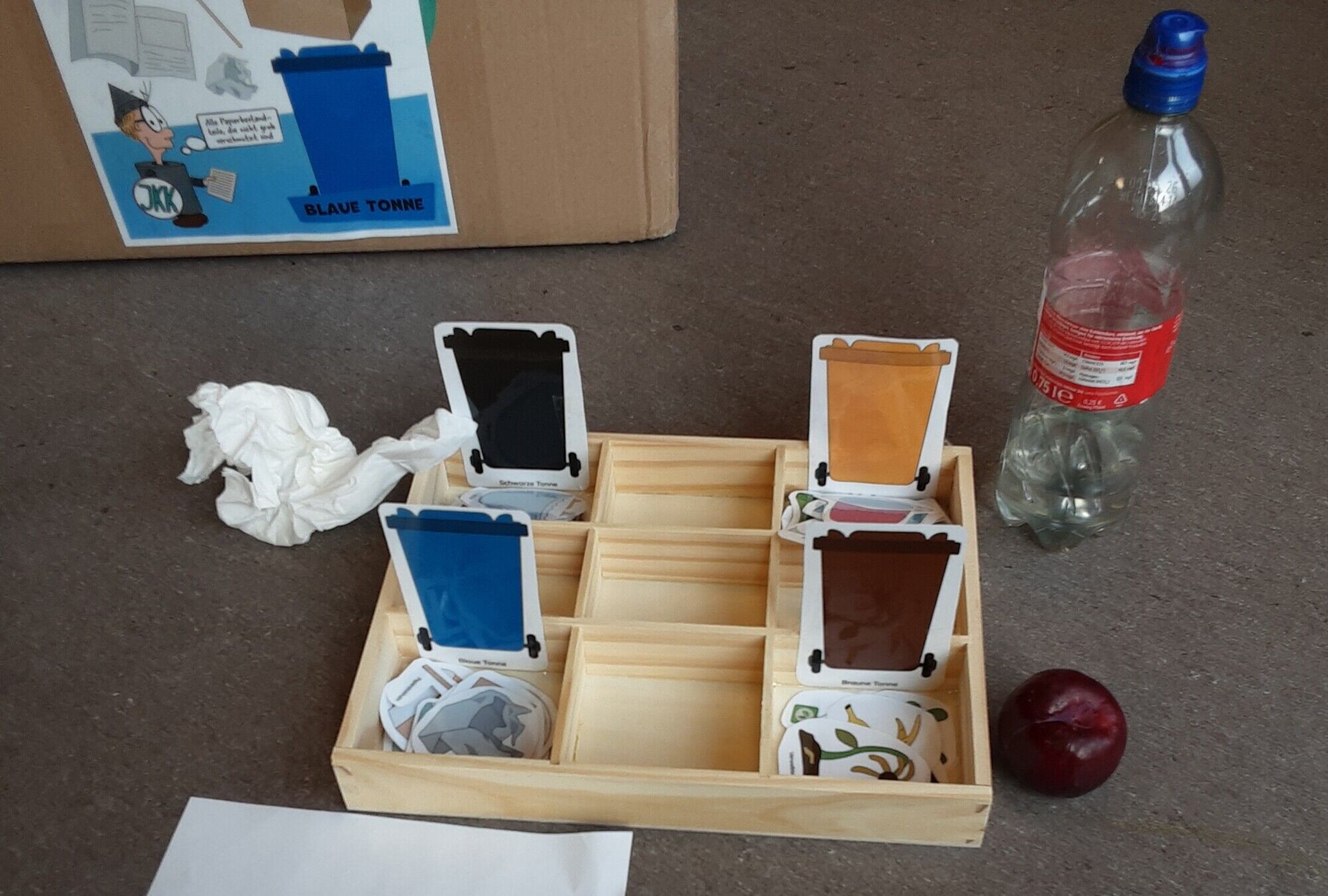 Eine Holzkiste mit 9 F&auml;chern in denen Bilder von farbigen M&uuml;lltonnen einsortiert sind. Daneben Bilder von unterschiedlichen Arten von Abfall, welche den M&uuml;lltonnen zusortiert werden sollen.