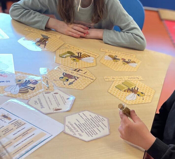 Karten mit Text, Bienen und Waben liegen auf einem Tisch um den Kinder sitzen