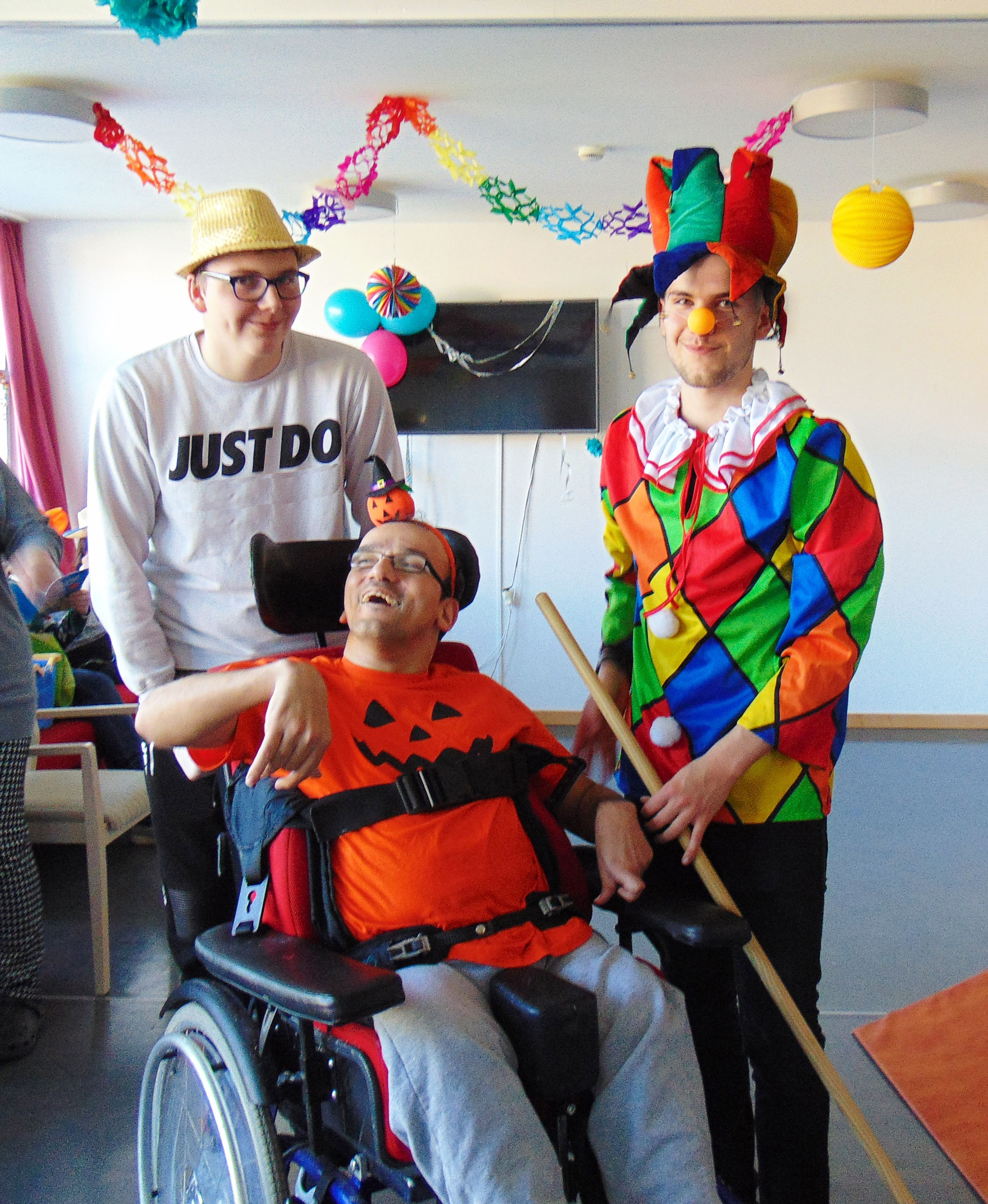 Zwei Menschen stehen im Kostüm hinter einem lachenden Bewohner im Rollstuhl.