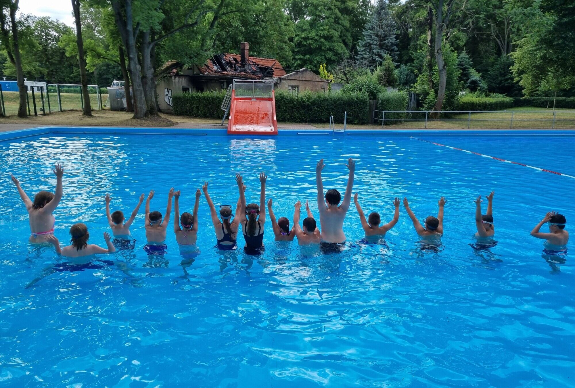 Viele Kinder sind in einem großen Schwimmbecken in einer Reihe stehend von hinten zu sehen. Sie strecken die Arme in die Höhe.