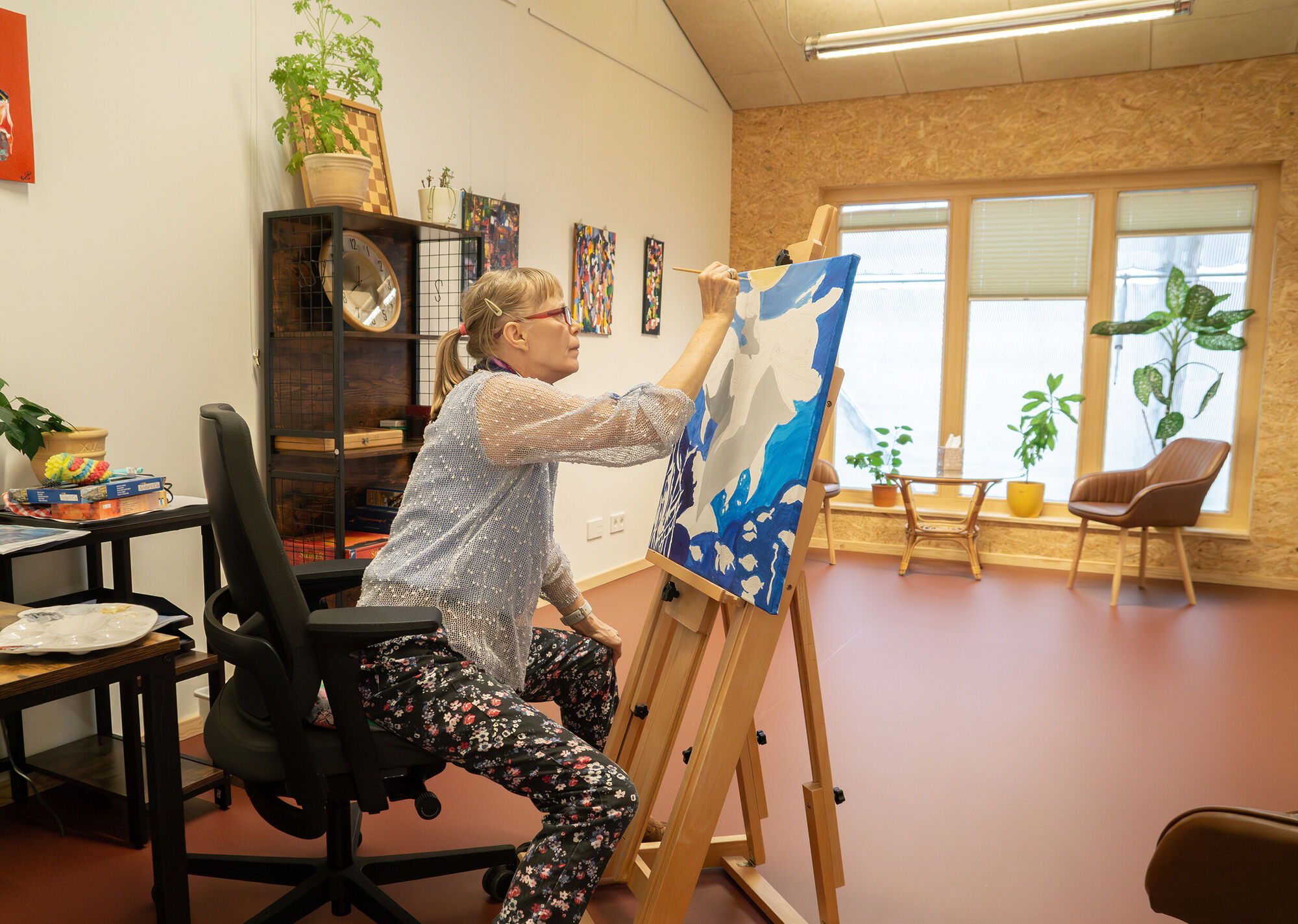 eine Frau sitzt in einem gemütlichen Raum vor einer Staffelei und malt mit Farben ein Bild