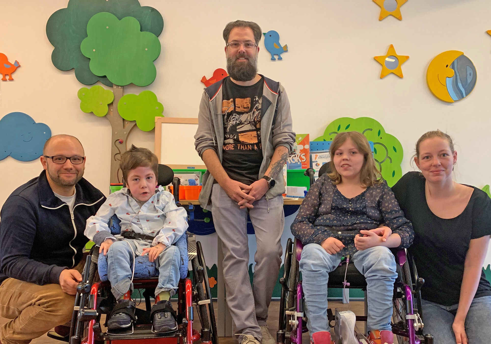 Zwei Kinder im Rollstuhl lächeln gemeinsam mit drei Erwachsenen in die Kamera.