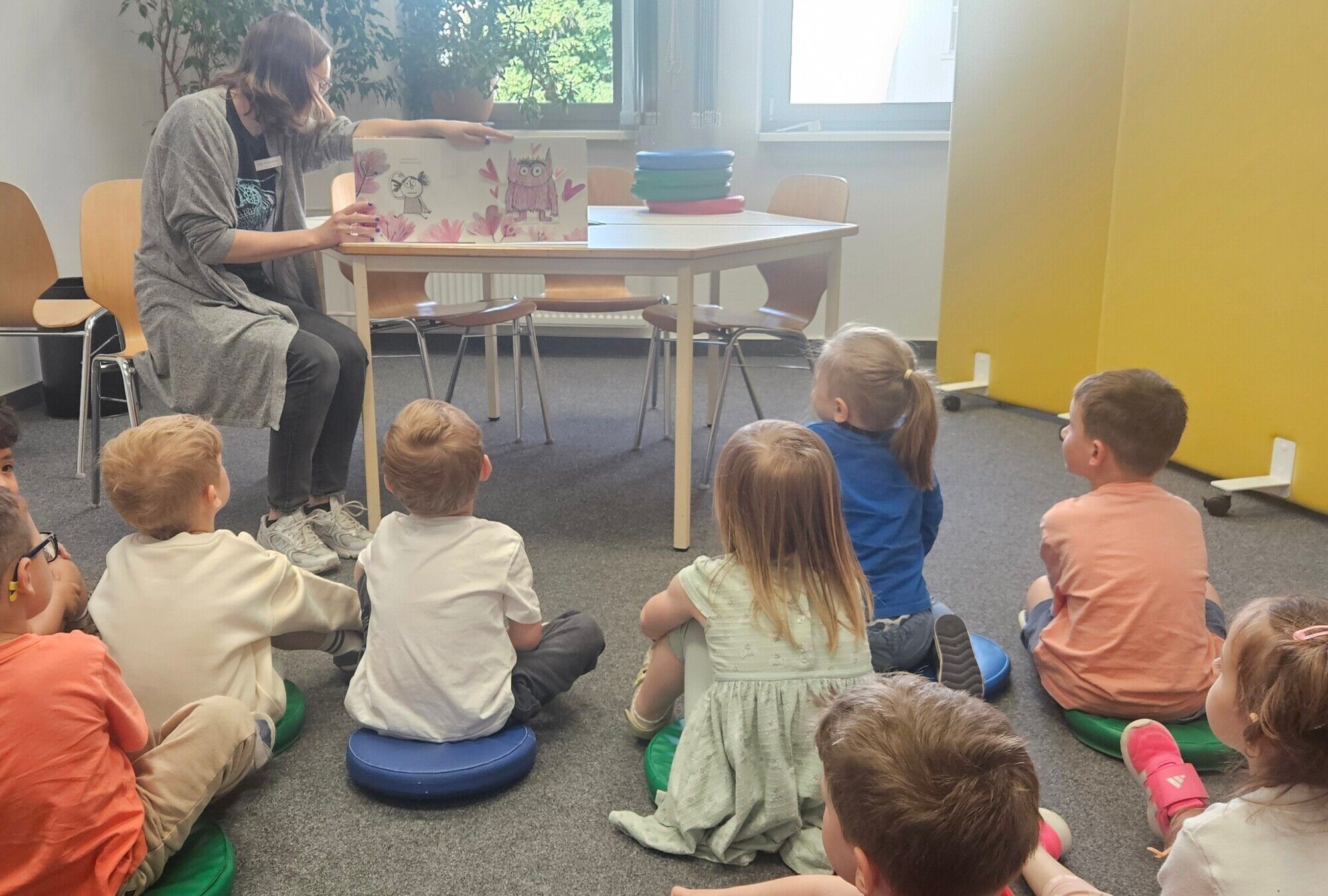 Kinder sitzen auf dem Boden und h&ouml;ren einer Frau beim Vorlesen zu. Gemeinsam schauen sie Bilder an