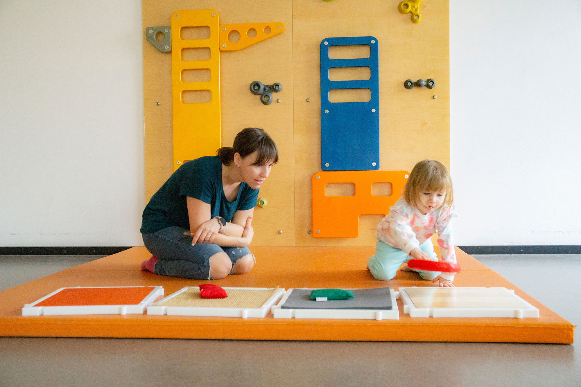 Eine terapeutin sitz mit einem Kind auf orangenen Bodenmatten. Das Kind sortiert große Figuren.