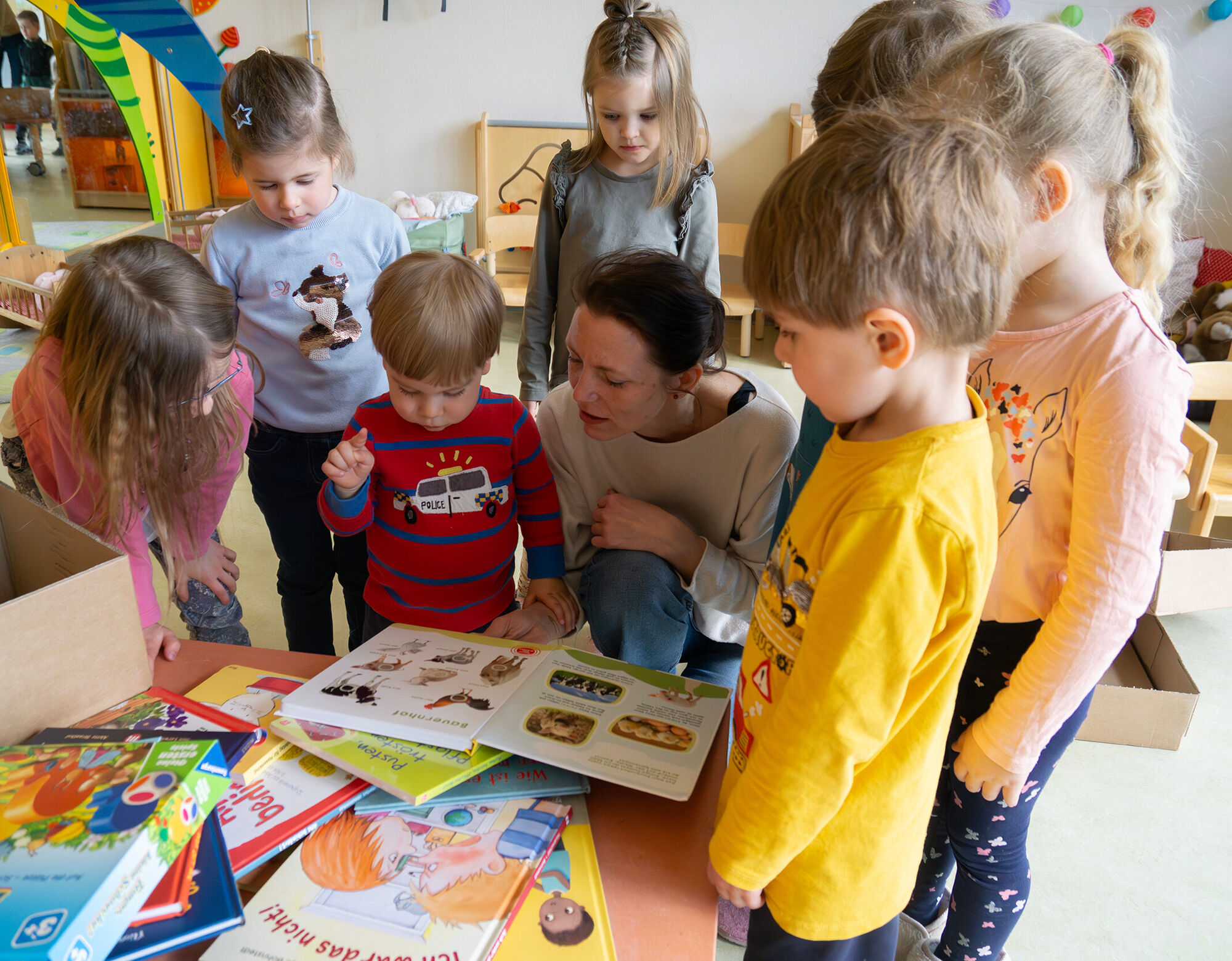 Kinder stehen und hocken vor einem Tisch mit vielen B&uuml;chern. Eine Erzieherin schaut sie mit Ihnen gemeinsam an