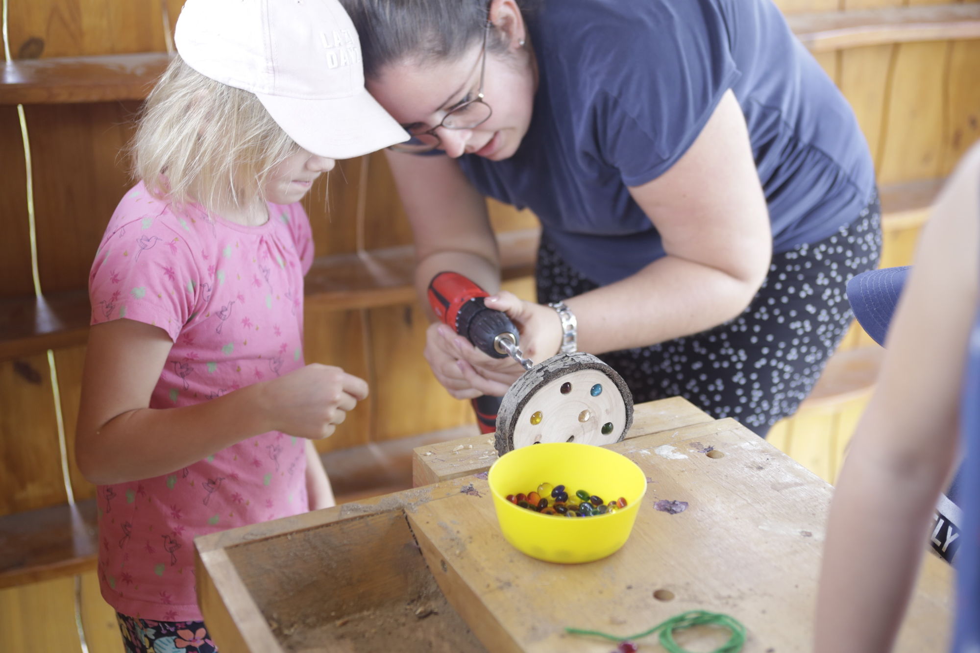 Eine Erzieherin zeigt einem Kind wie mit einer Bohrmaschine Löcher in eine Holzscheiben gebohrt werden. In einigen Löchern befinden sich bunte Glassteine