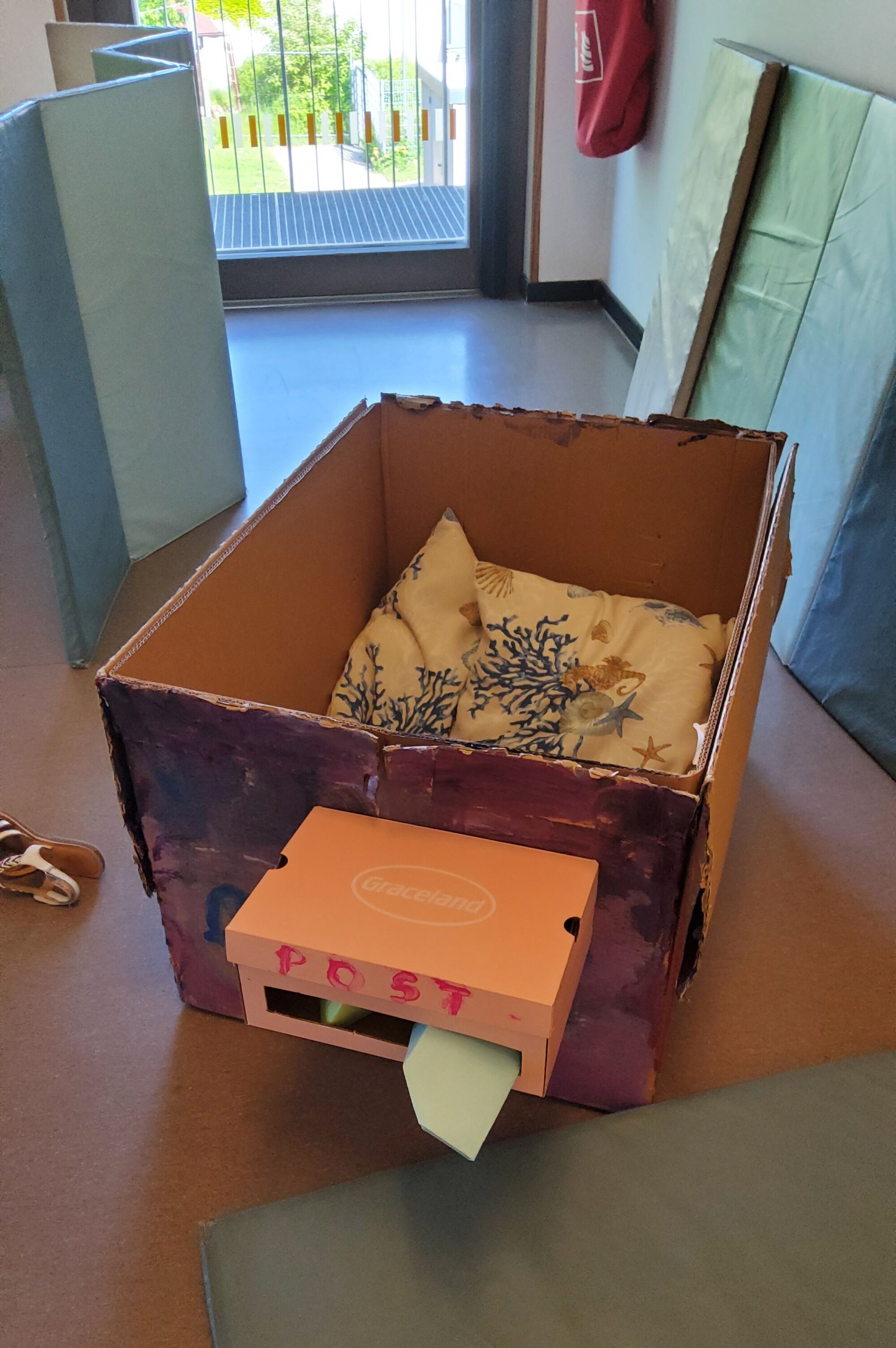 Ein gro&szlig;er Karton mit einem Kissen drin und einem gebastelten Postkasten an einer Seite