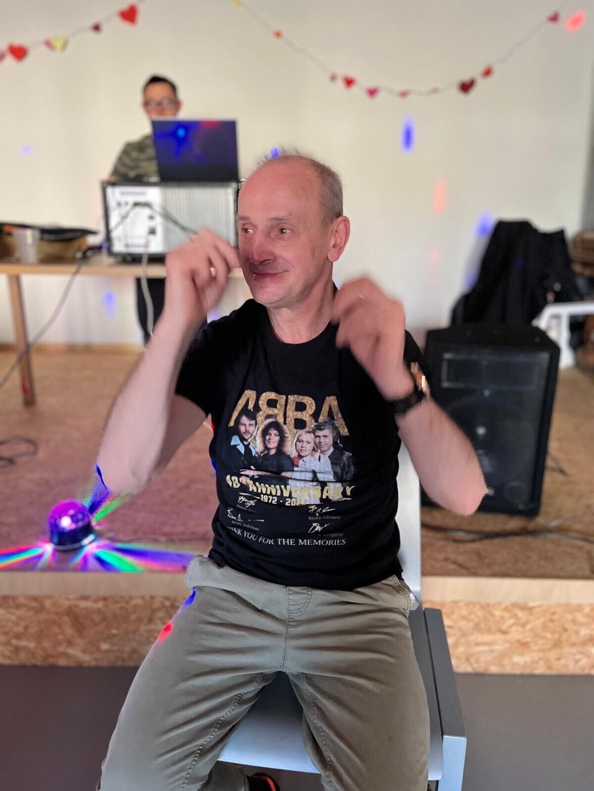 Ein Mann sitz l&auml;chelnd auf einem Stuhl und hebt tanzend die Arme