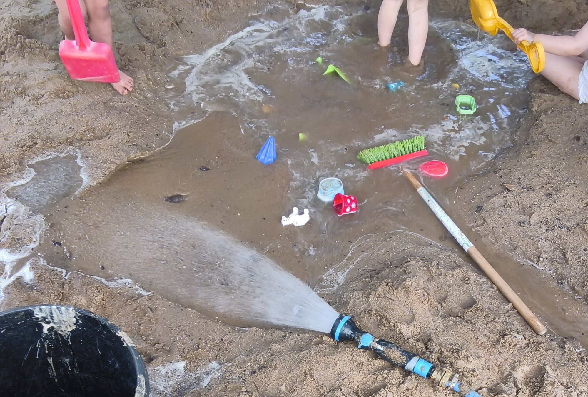 Eine gro&szlig;e Pf&uuml;tze im Sand, darin Kinder in Windeln, Sandspielzeug und ein Gartenschlauch