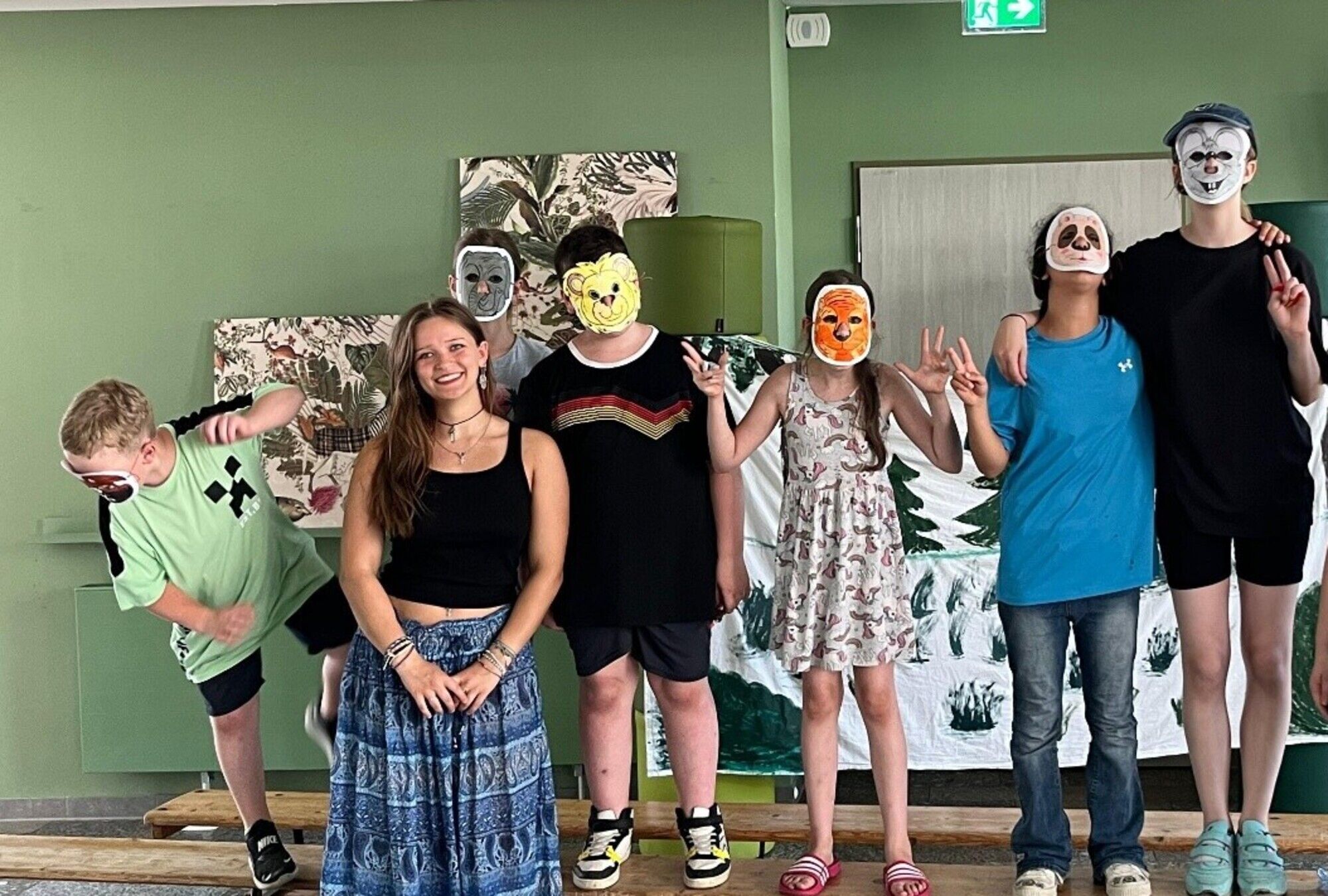 Eine junge Frau blickt lachend in die Kamera. Hinter Ihr Kinder mit selbstgebastelten Masken vor dem Gesicht.