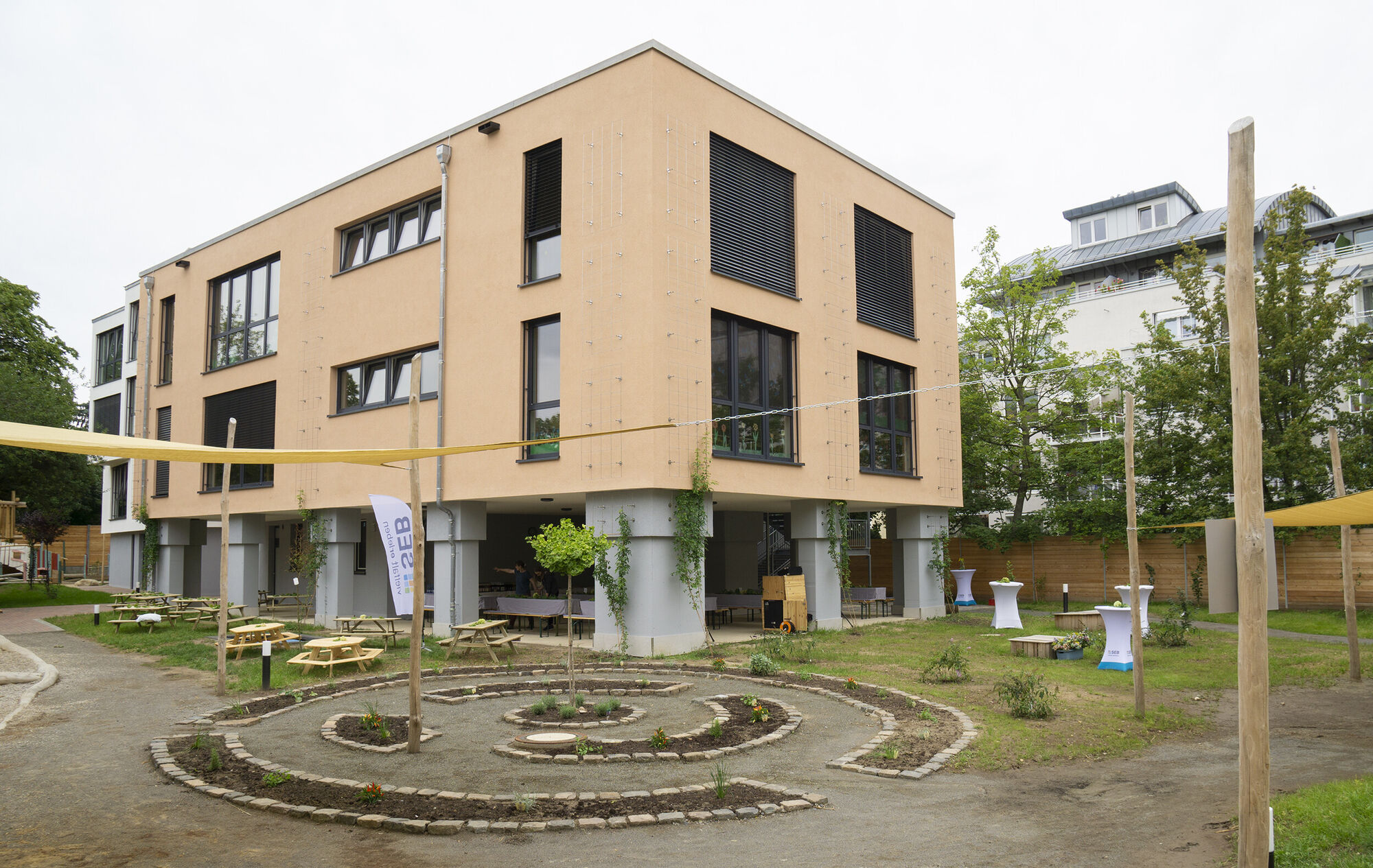 ein großes Gebäude mit vielen Fenstern. Davor sind Beete in einem Rundell angelegt.