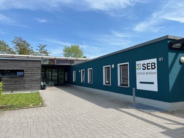 Der Eingangsbereich zu einem flachen Gebäude mit Holz- und petrolfarbener Putzfassade. An einer Ecke ist das SEB-Logo zu sehen.