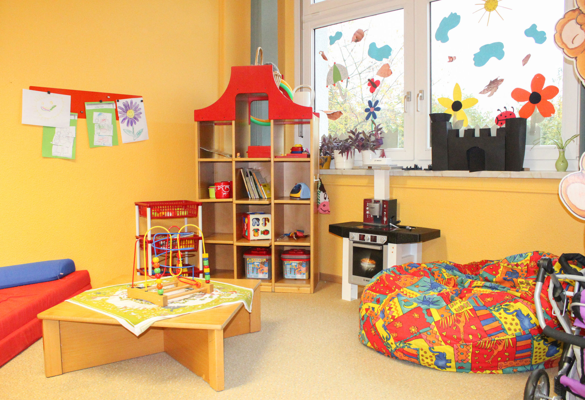 Ein Raum mit Kinderküche, Regal mit Spielen und einem großen bunten Sitzkissen.