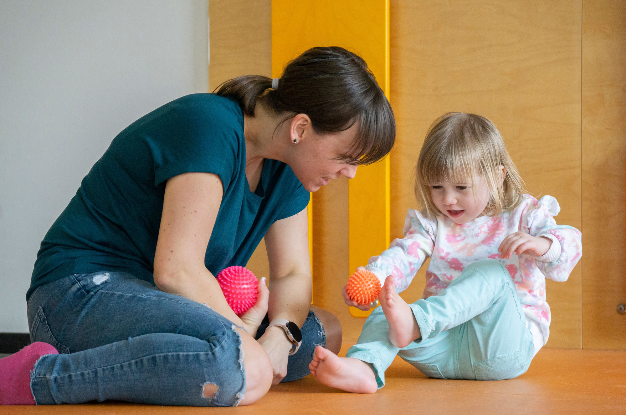 Eine Therapeutin lächelt einem Kind zu, welches einen Stacheligen Ball an seine nackten Füße hält.