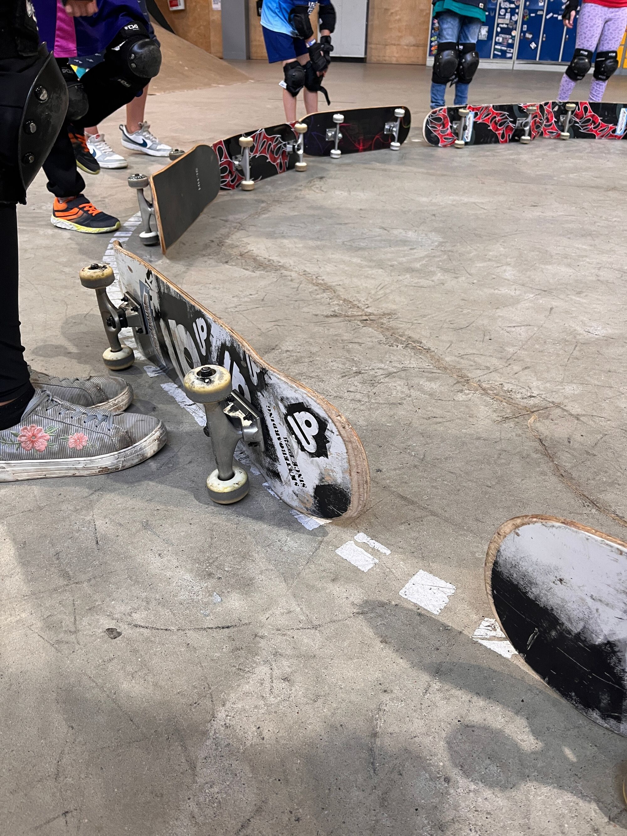 Skateboards liegen an einer Linie, dahinter sind Beine und Füße mit Knieschützern zu sehen.