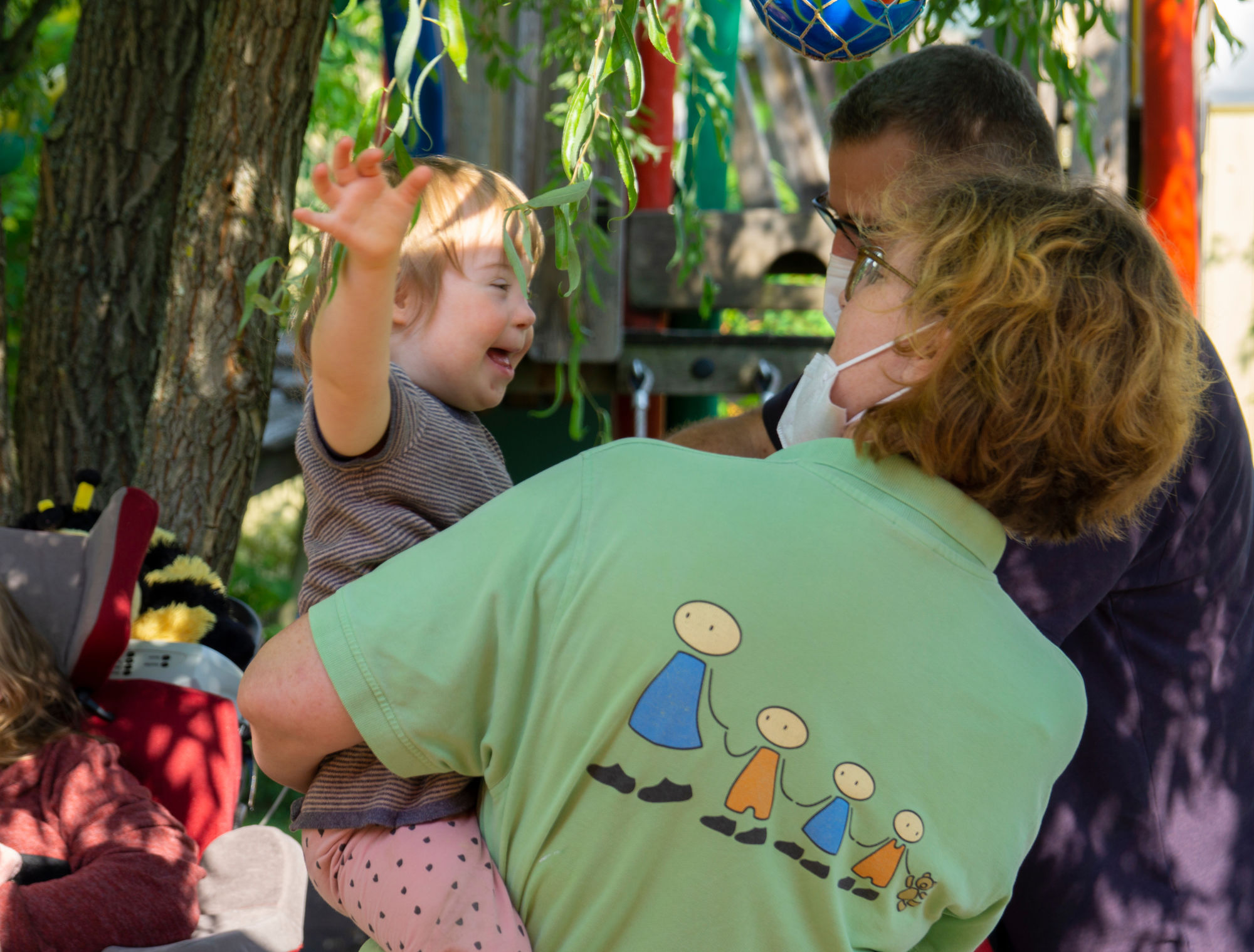 Eine Frau in grünem T-Shirt hat ein kleines Mädchen auf dem Arm, das die Hand in die Höhe streckt und lacht.