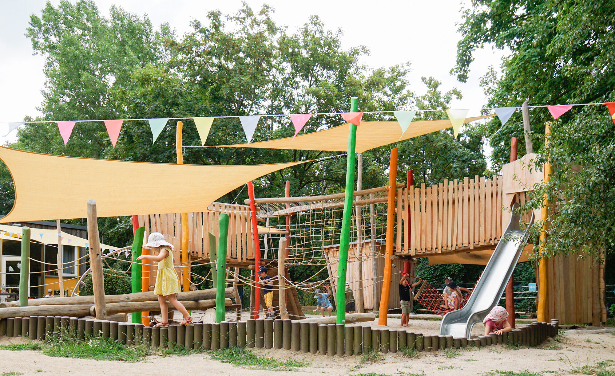 Kinder spielen in einem Garten mit großer bunter Kletter- und Balancierstrecke, Rutsche, Sandkästen und Wiese.