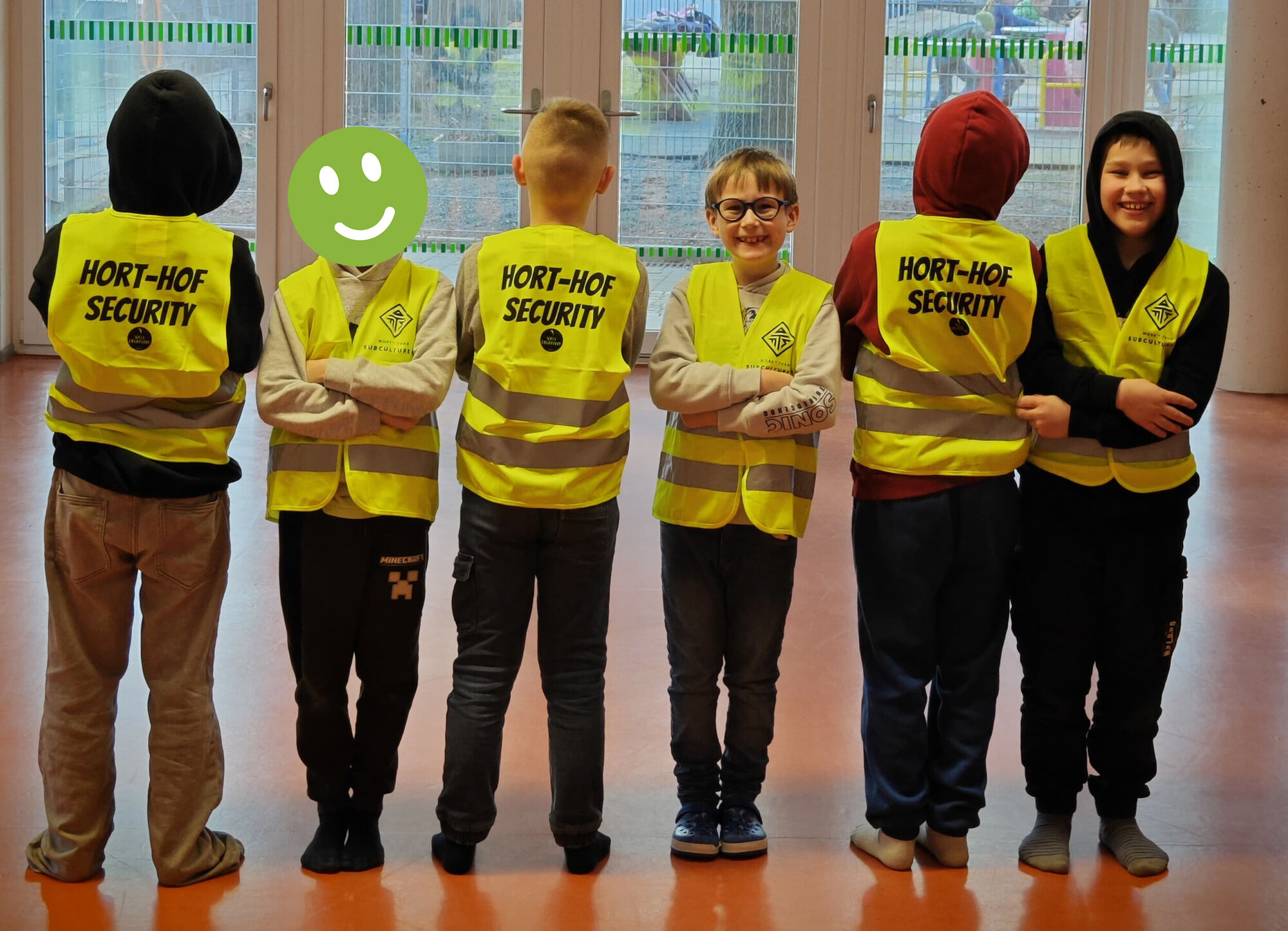 Kinder stehen in einer Reihe und tragen Westen mit der Aufschrift "Hort-Hof-Security" und lachen in die Kamera
