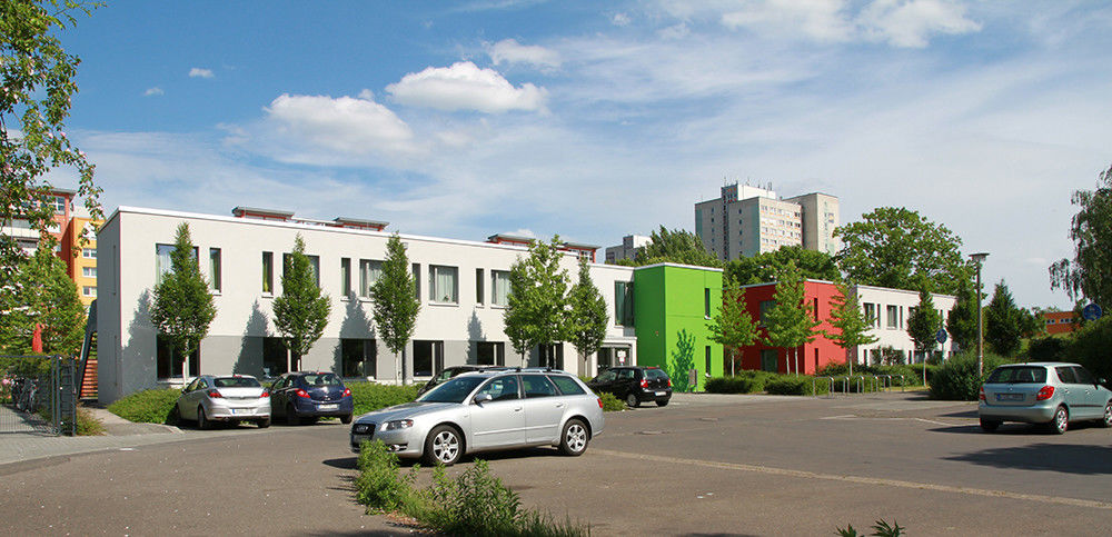 Ein längliches helles gebäude mit roten und grünen Gebäudeteilen. Davor stehen Autos auf Parkplätzen