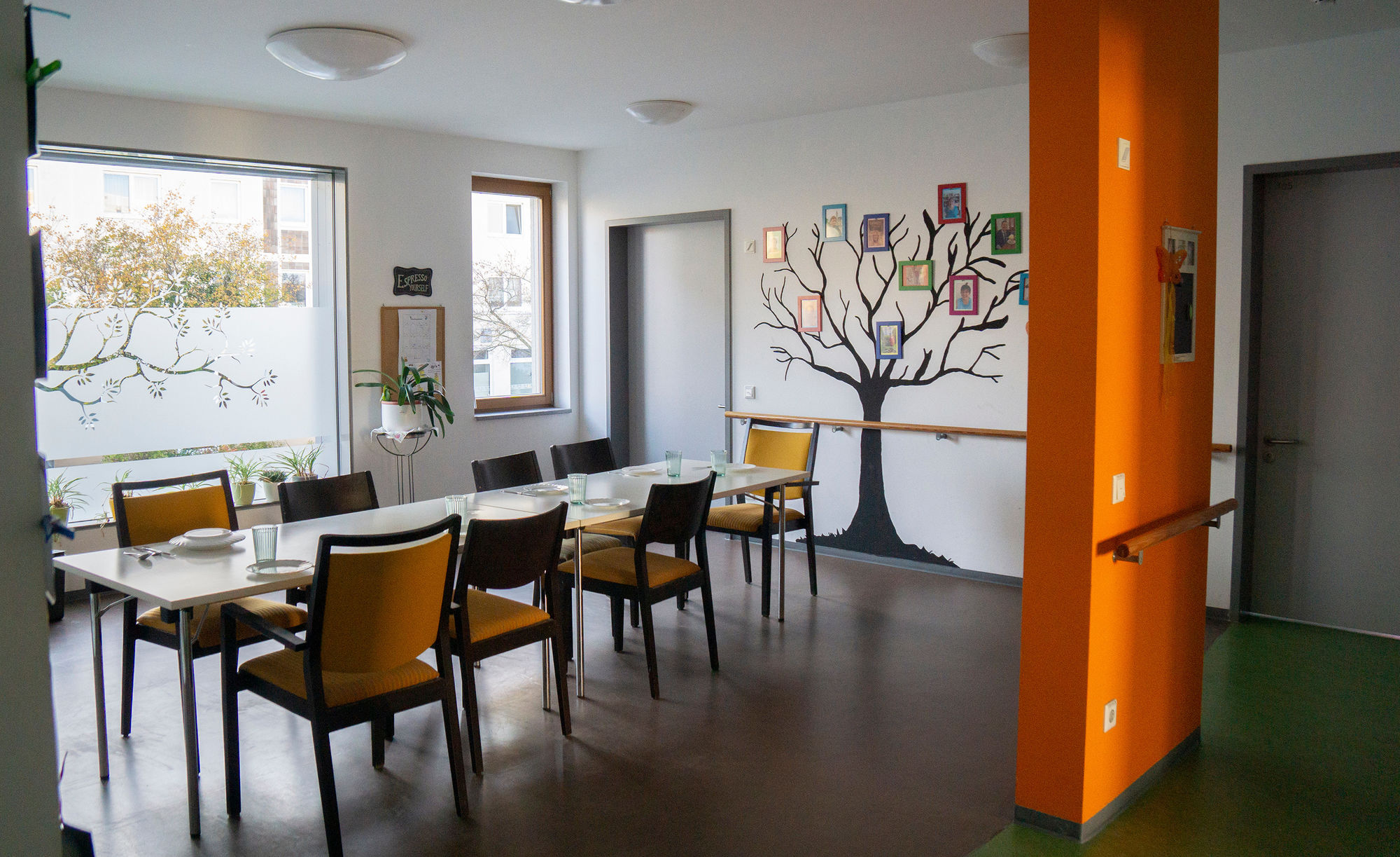 Ein Gemeinschaftsbereich mit Tisch und Stühlen, großen Fenstern und einem Baum mit Bildern an der Wand.