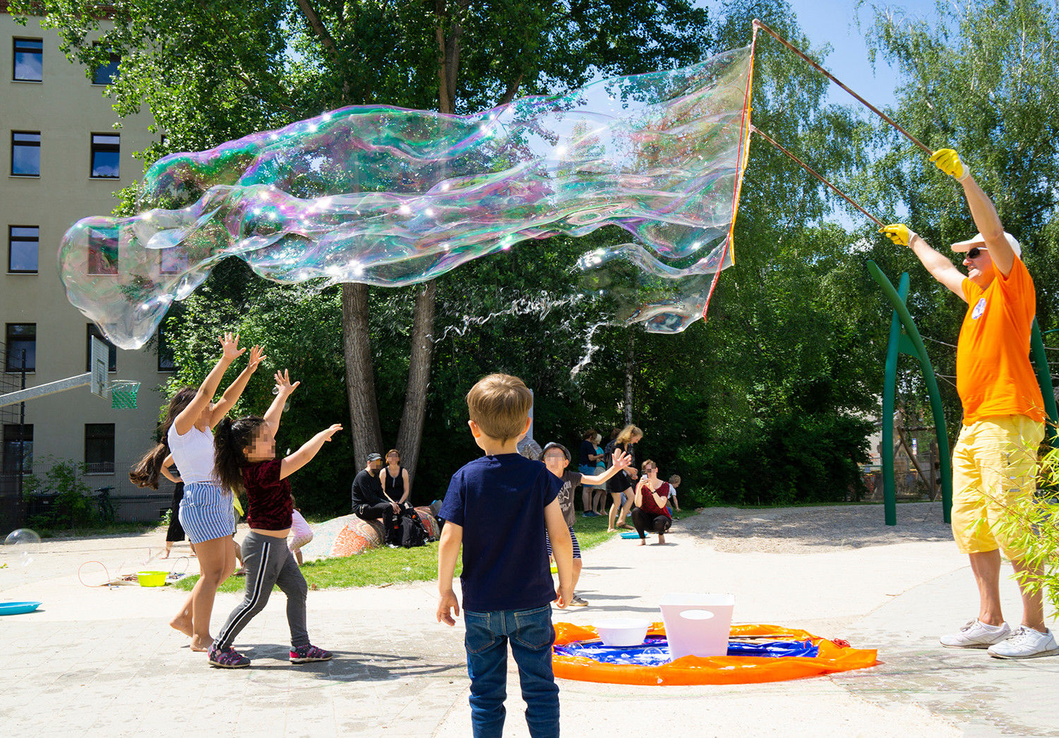 Ein Mann macht eine riesige Seifenblase, die in der Sonne schimmert. Drumherum sind Kinder, die versuchen sie platzen zu lassen