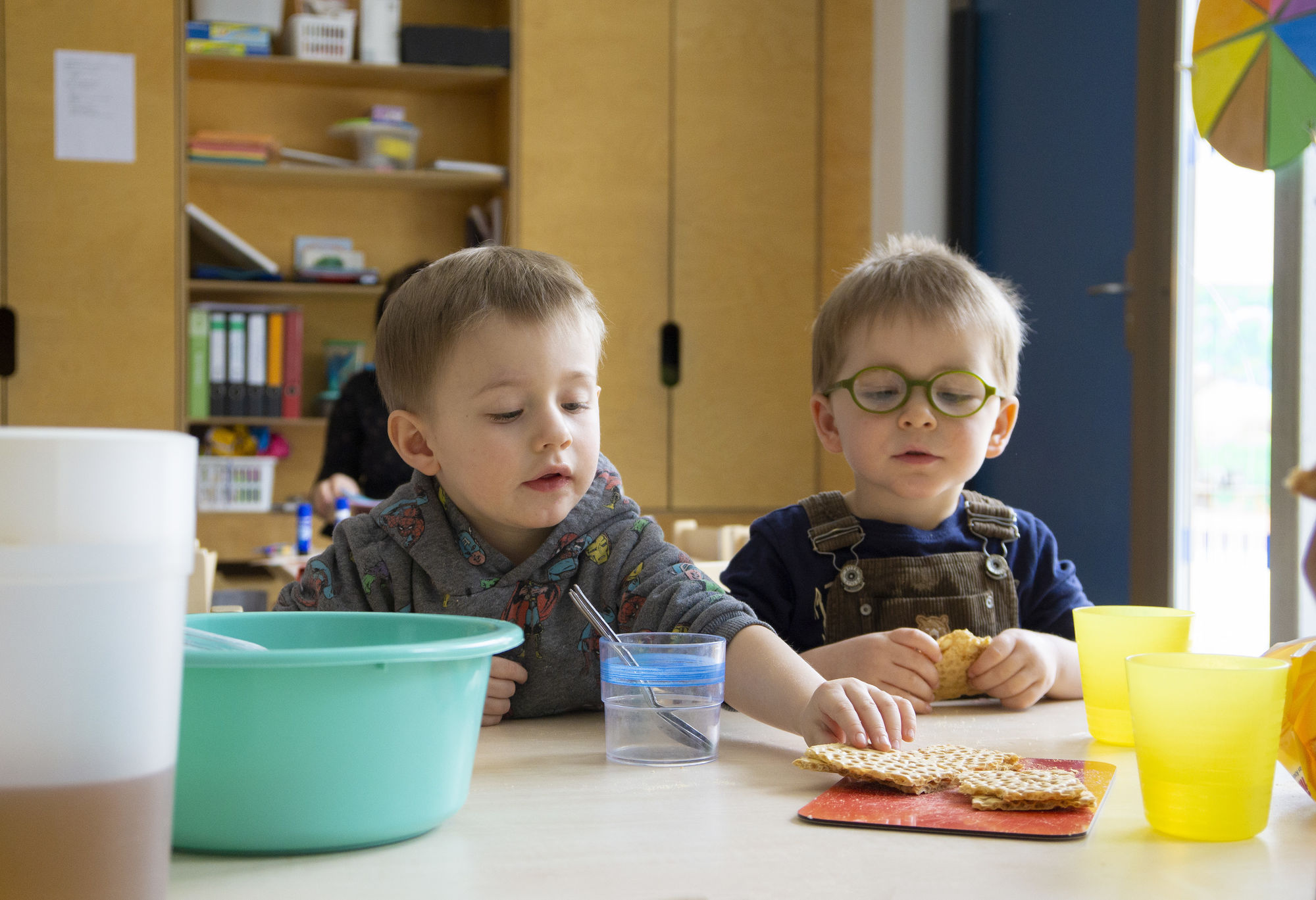 Zwei Kinder sitzen an einem Tisch mit Trinkbechern und nehmen sich Knäckebrot von einem Teller