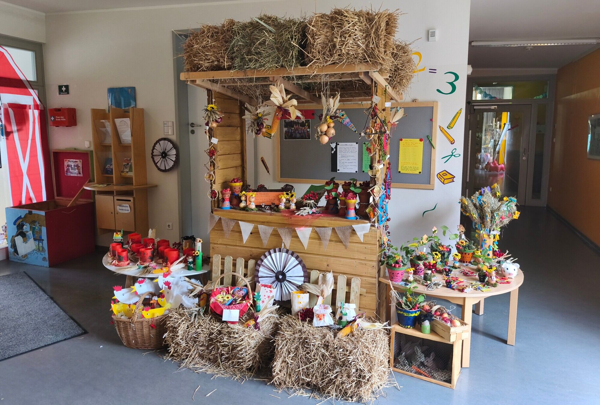 Ein Verkaufsstand aus hellem Holz, dekoriert mit großen Strohballen und vielen bunten gebastelten Bauernmarktartikeln