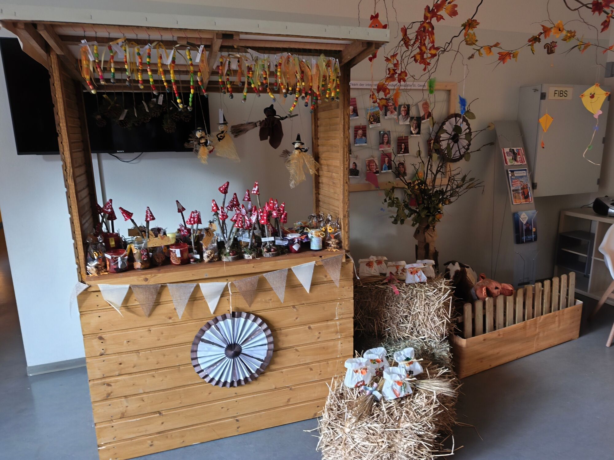 Ein Verkaufsstand aus hellem Holz, dekoriert mit großen Strohballen und vielen bunten gebastelten Bauernmarktartikeln