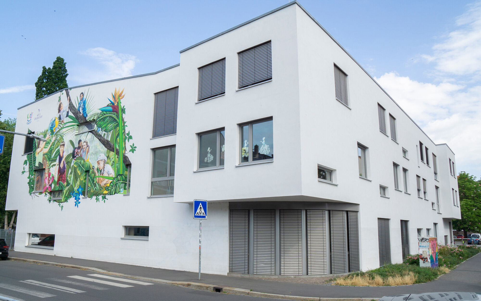 Ein weißes Gebäude mit einem großen Gaffitti, auf welchem Kinder, Tiere und Dschungelpflanzen zu sehen sind.