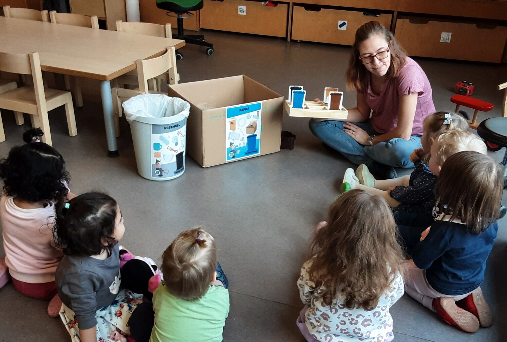 Eine Holzkiste mit 9 F&auml;chern in denen Bilder von farbigen M&uuml;lltonnen einsortiert sind. Daneben Bilder von unterschiedlichen Arten von Abfall, welche den M&uuml;lltonnen zusortiert werden sollen.