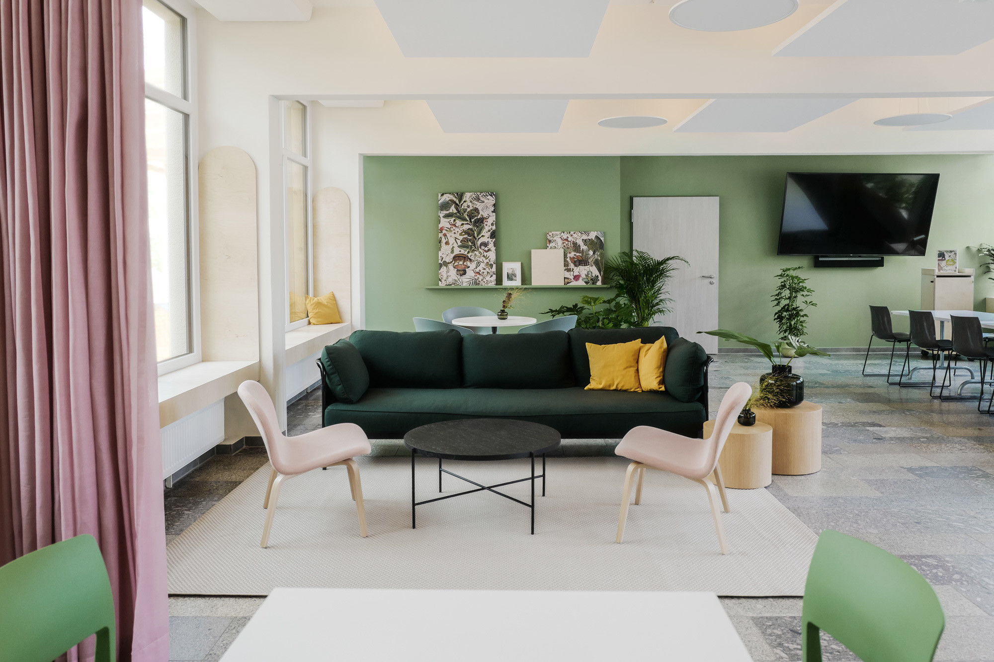 © loeserbettels Eim großer heller Raum mit einer Loungeecke mit grünem Sofa und gepolsterten rosa Stühlen, einer langen Tafeln mit vielen Stühlen und einem großen Bildschirm an der Wand.