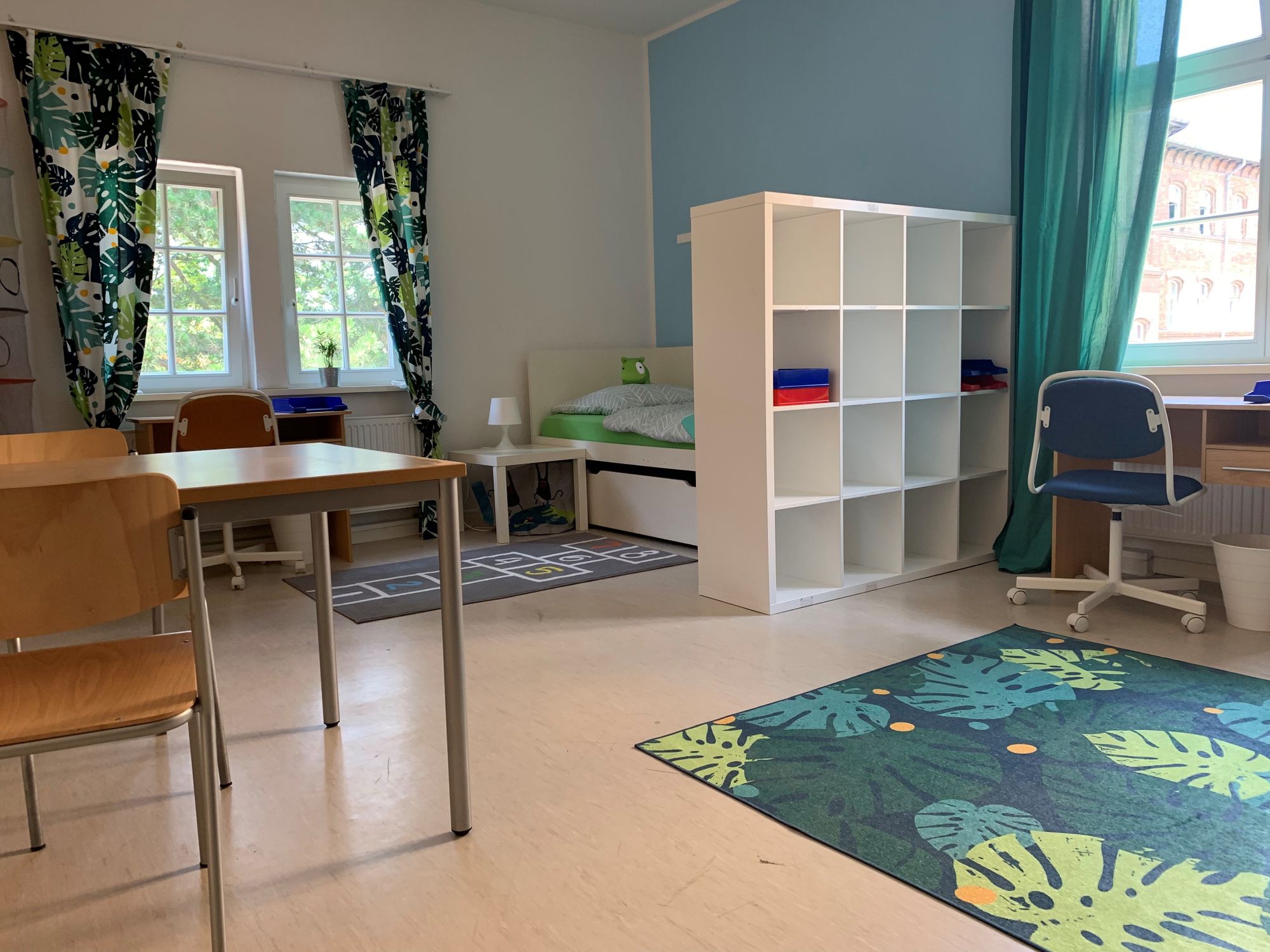 Ein fröhliches Zimmer mit Schreibtisch, Bett, Tisch und Stühlen bunten Vorhängen und Teppich.