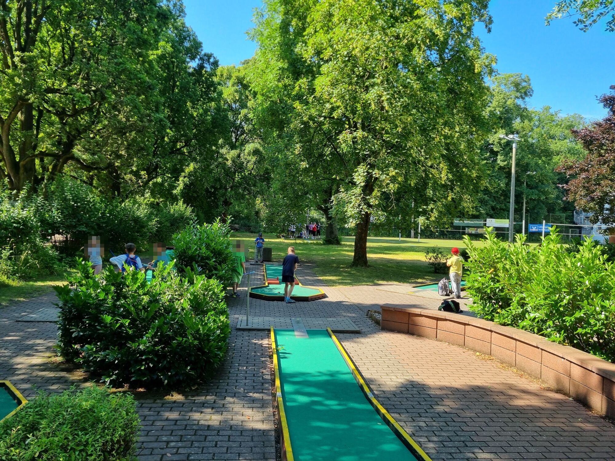 Kinder spielen Minigolf im freien zwischen vielen Bäumen bei strahlendem Sonnenschein.