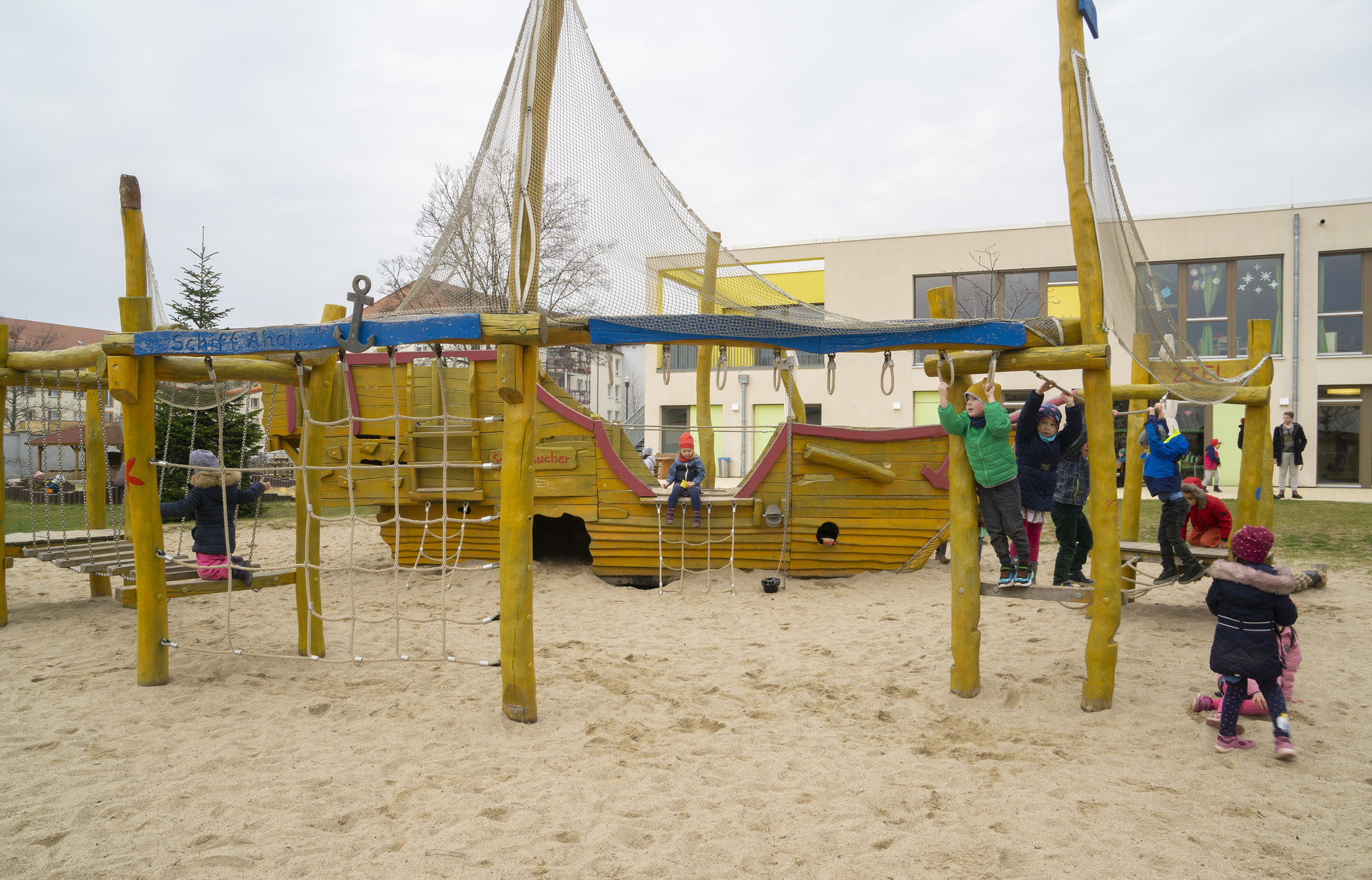 Kinder klettern auf Holzbalken und an Netzen und hangeln sich an Griffen entlang. Im Hintergrund ist das Kletterpiratenschiff zu sehen.