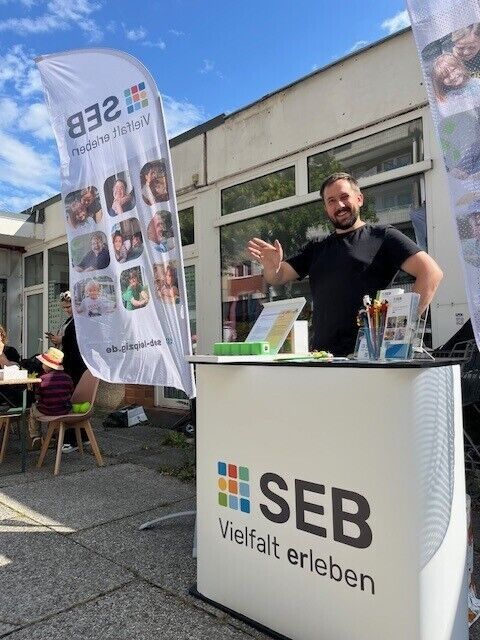 Vor einem Gebäude ist ein SEB-Infostand aufgebaut. Ein Mitarbeiter steht dahinter, lächelt und winkt in die Kamera