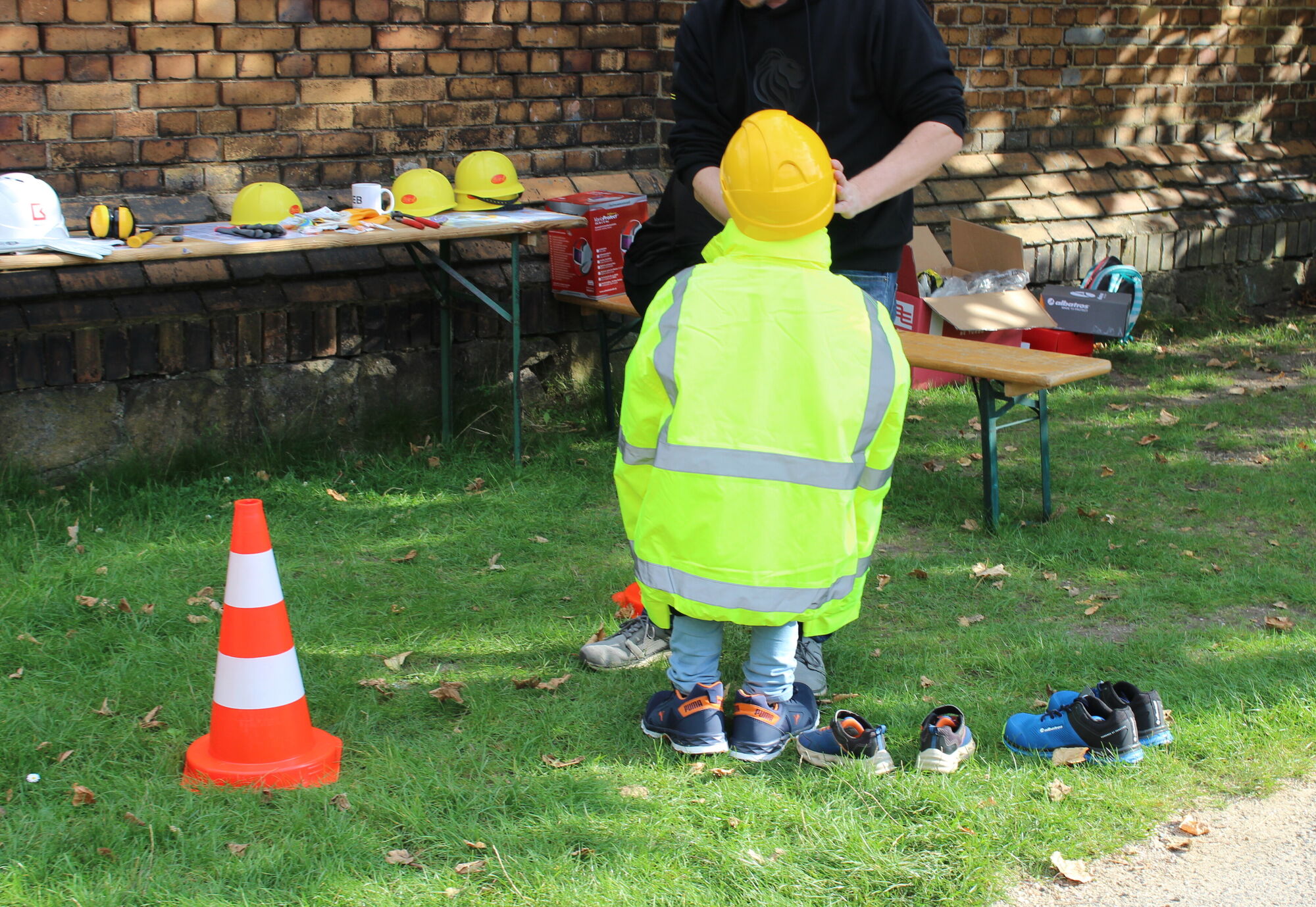 Ein Kind mit Helm und gro&szlig;er gelber Warnweste an einem Aktionsstand mit Werkzeugen, betreut durch einen Erwachsenen