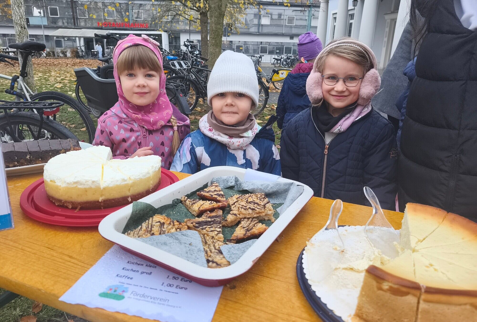 Drei Kita-Kinder stehen dick angezogen hinter einem Tisch und verkaufen Kuchen