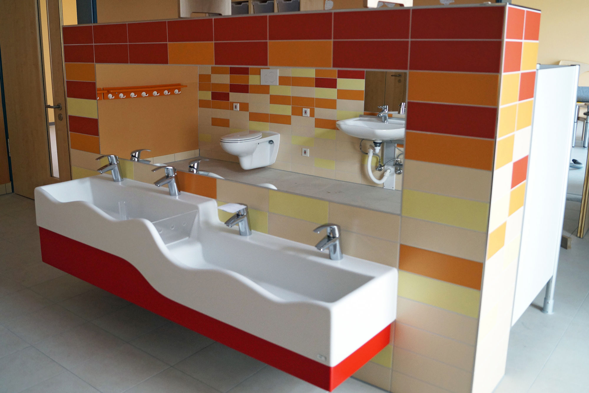 Eine bunt geflieste Wand mit einem breiten Waschbecken und Spiegeln im Kinderhöhe.