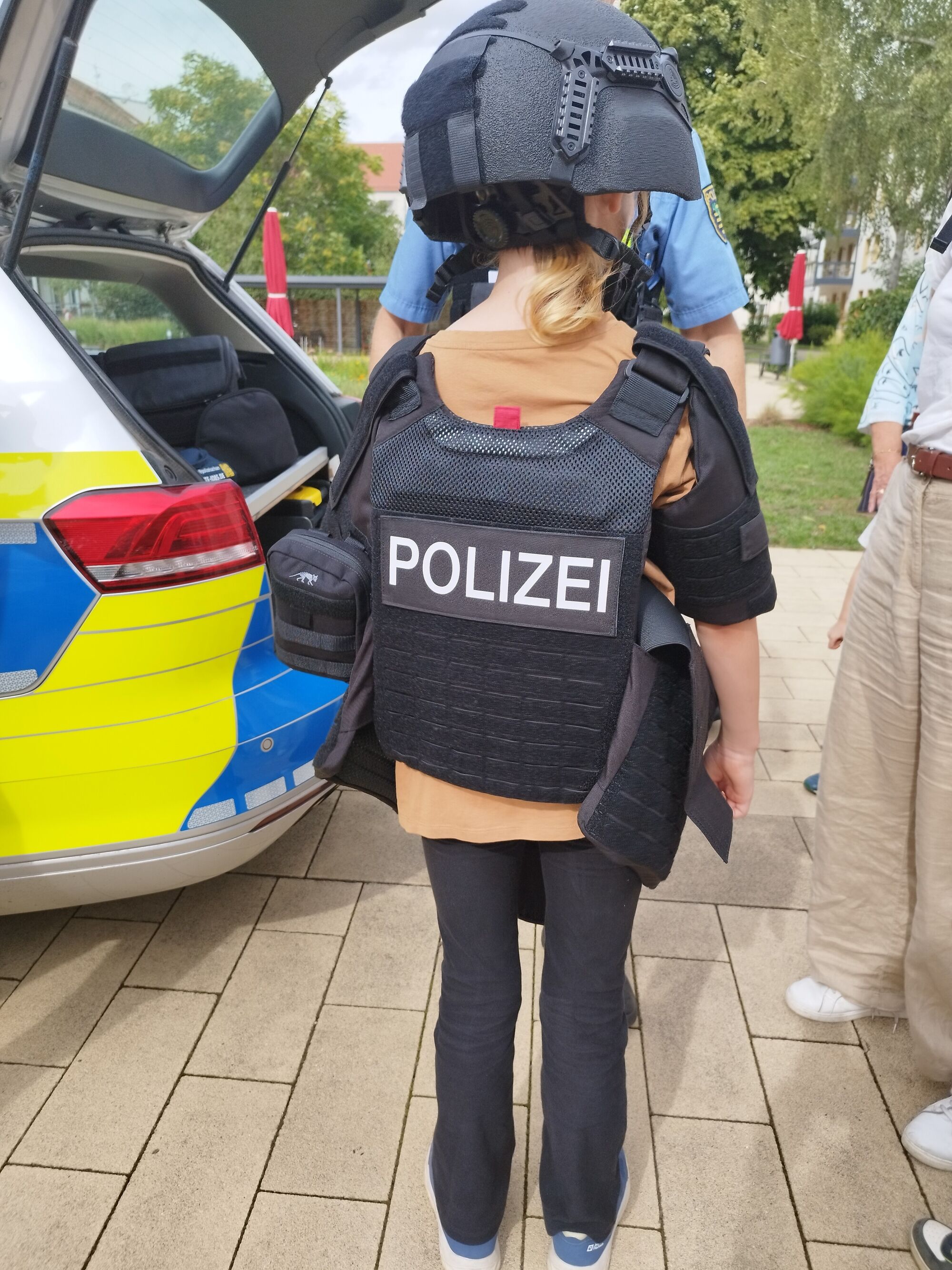 Ein Kind mit Polizeiweste und Helm vor einem Polizeiauto
