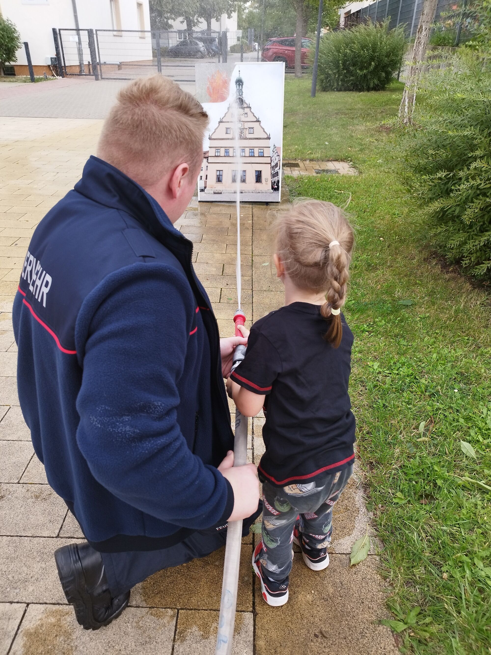 Ein Kind darf mit dem Feuerwehrschlauch auf ein gro&szlig;es Haus zielen, das auf eine Holzplatte gemalt ist und oben am Dach "brennt"